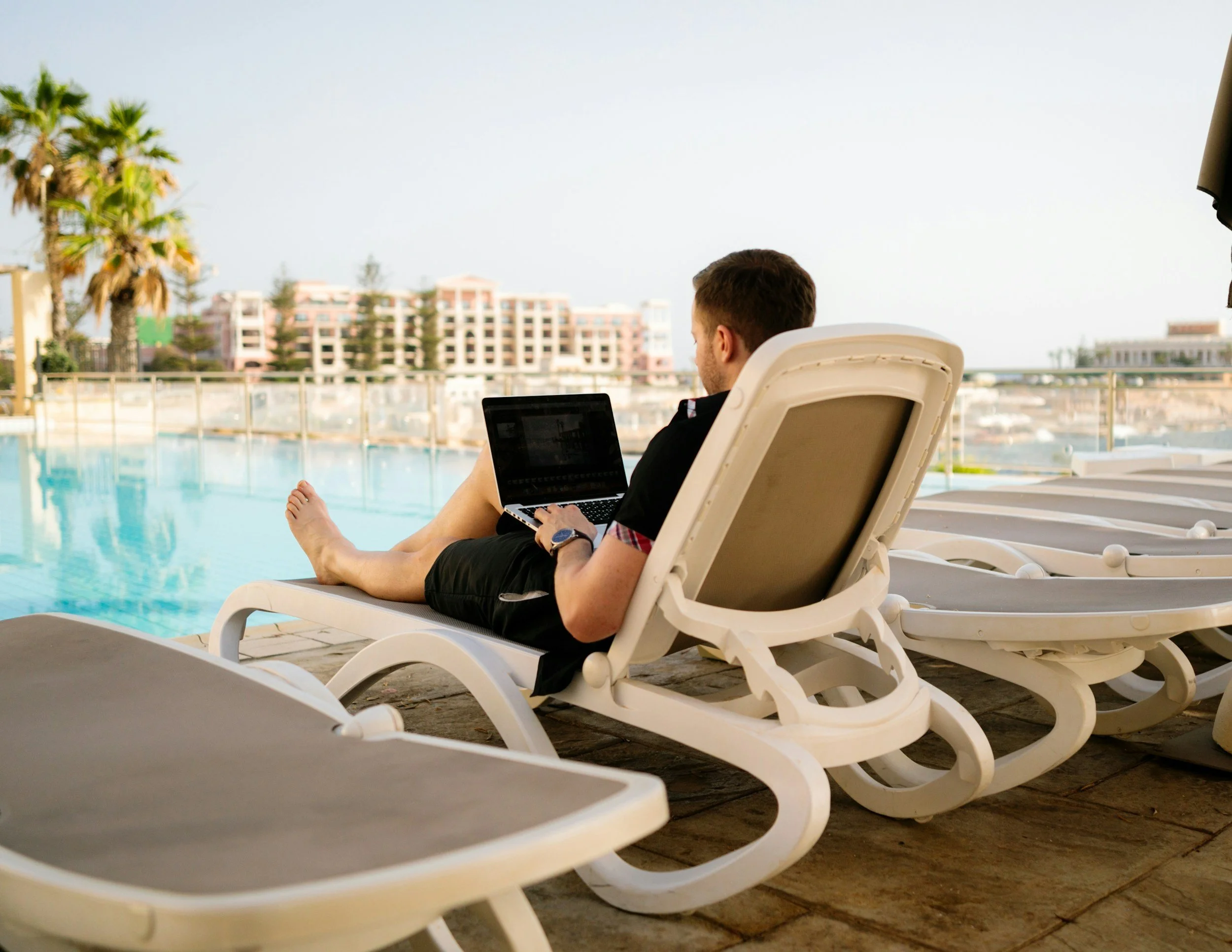 A man with a beard wearing a yellow t-shirt working on a laptop at a table outdoors with a river, bridge, and skyline with a mosque and other buildings in the background.