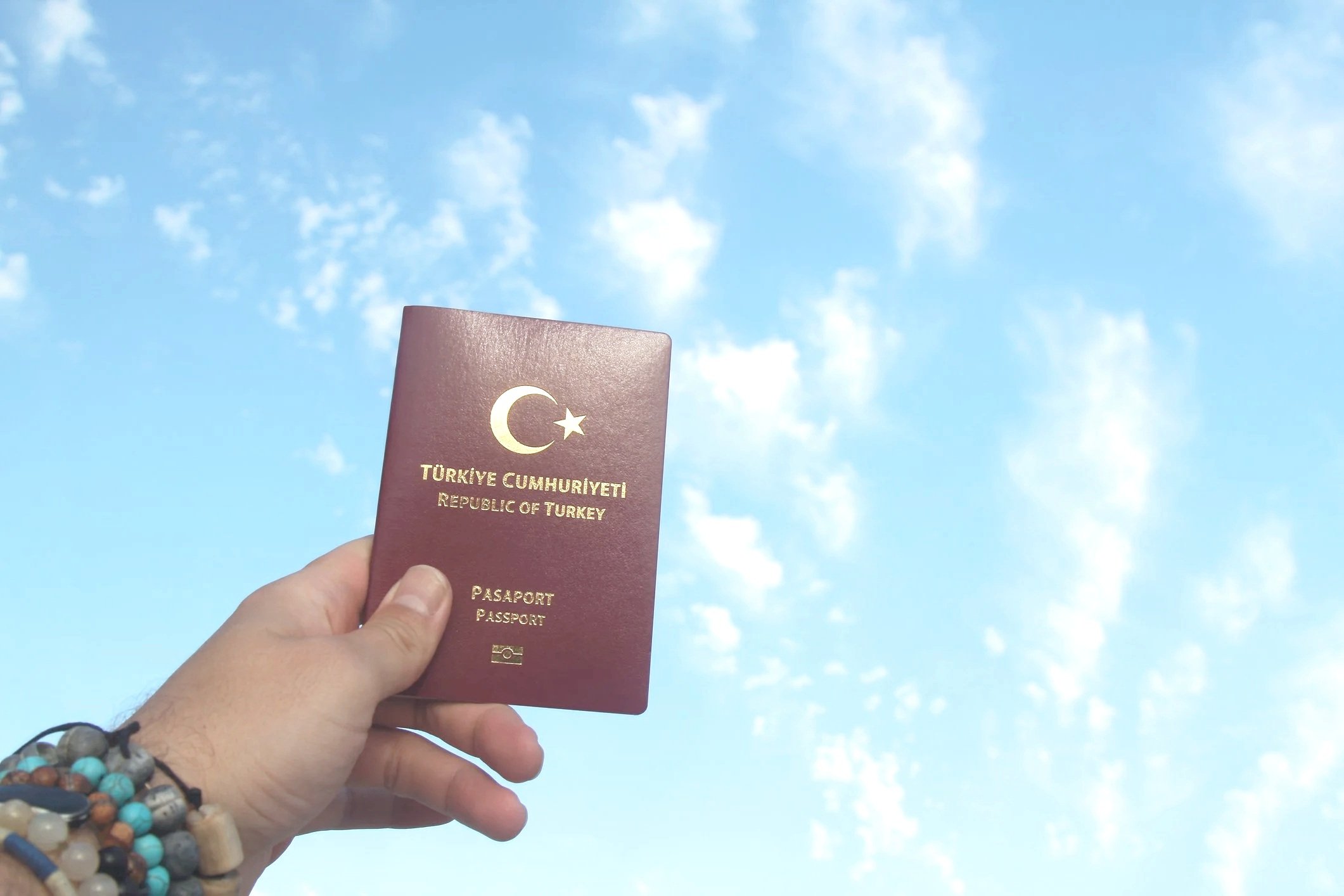 A man working at a desk with a computer, holding a passport, with a mobile phone showing a checkmark, a potted plant, a Turkish flag, and a bank building in the background.