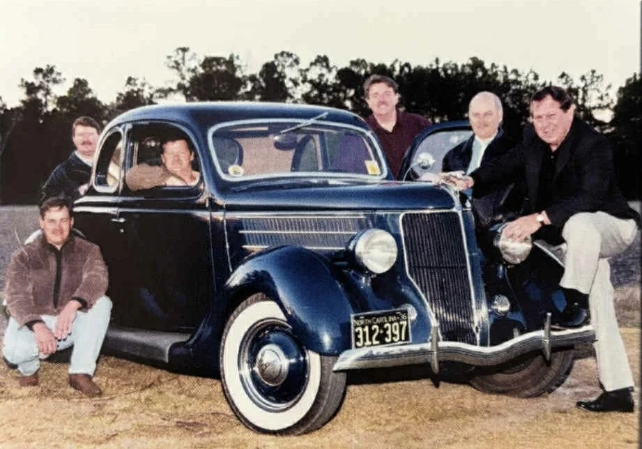 A vintage black car with white-walled tires, surrounded by six men outdoors during daytime, some sitting on the ground and others standing by the car, with trees in the background.
