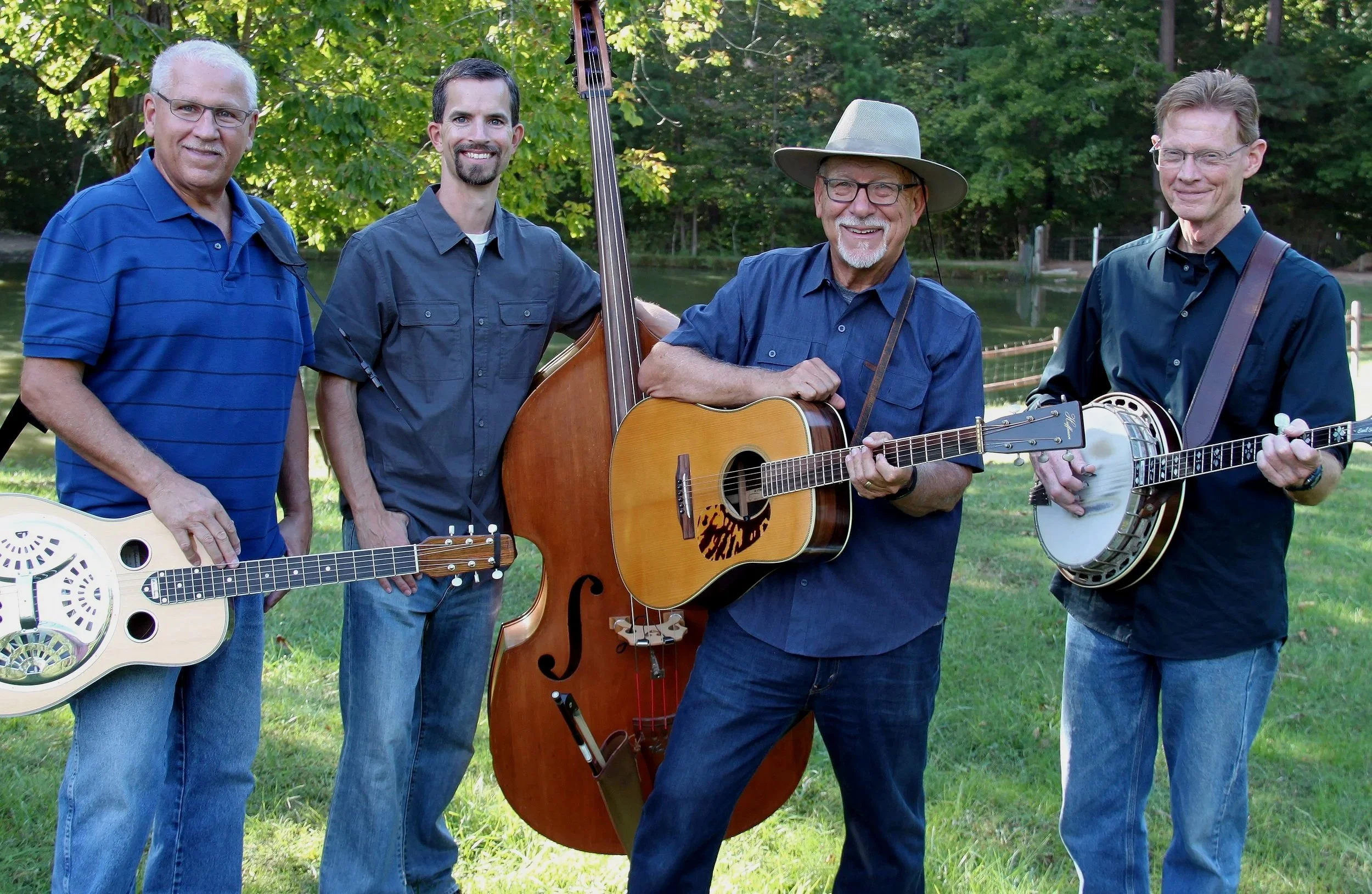 Four men standing outdoors in front of trees, each holding a musical instrument, smiling at the camera.