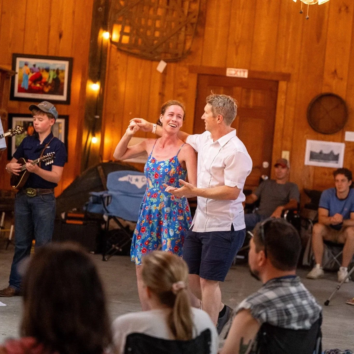A woman in a blue floral dress dancing with a man in a white shirt at an indoor gathering with wooden walls. A young boy is playing guitar nearby, and several people are watching or sitting in the background.