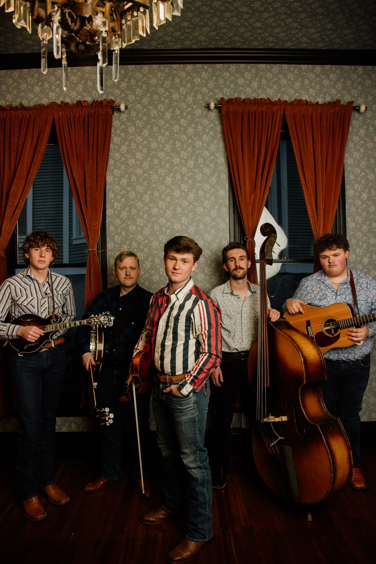 A group of five young musicians, Carson Peters and Iron Mountain, holding instruments, standing indoors in front of two windows with red curtains, with a chandelier hanging above.