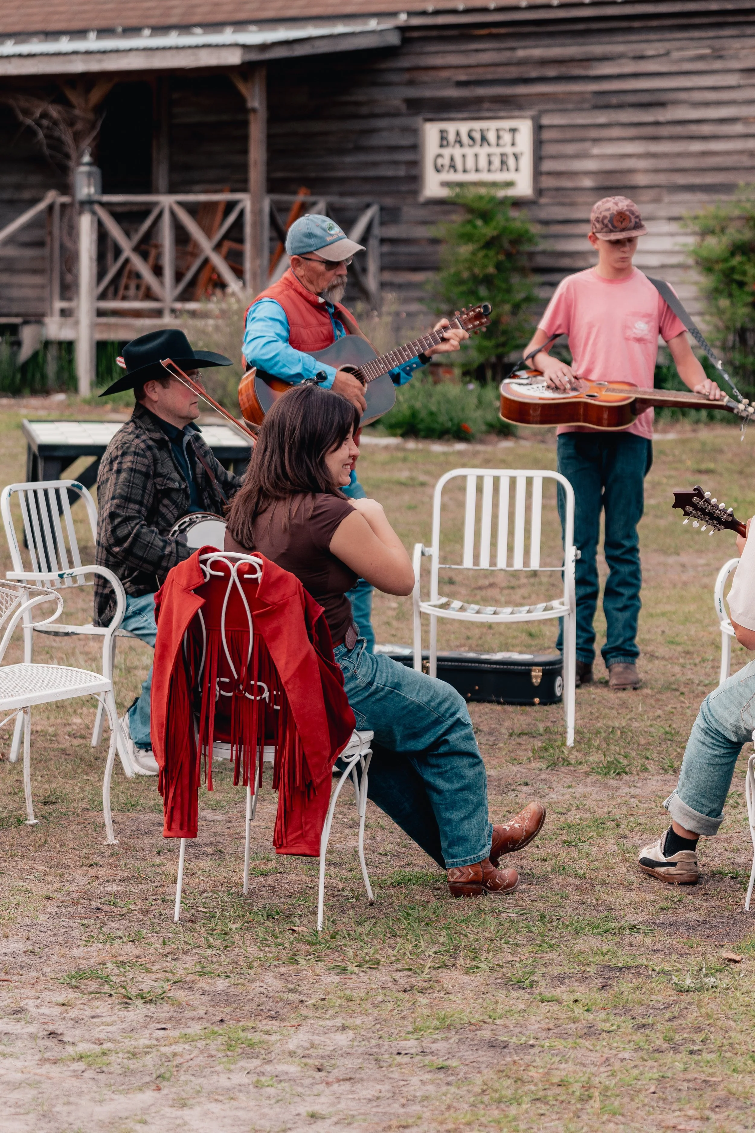 Group of people jamming outdoors, some playing guitars, sitting on white chairs.