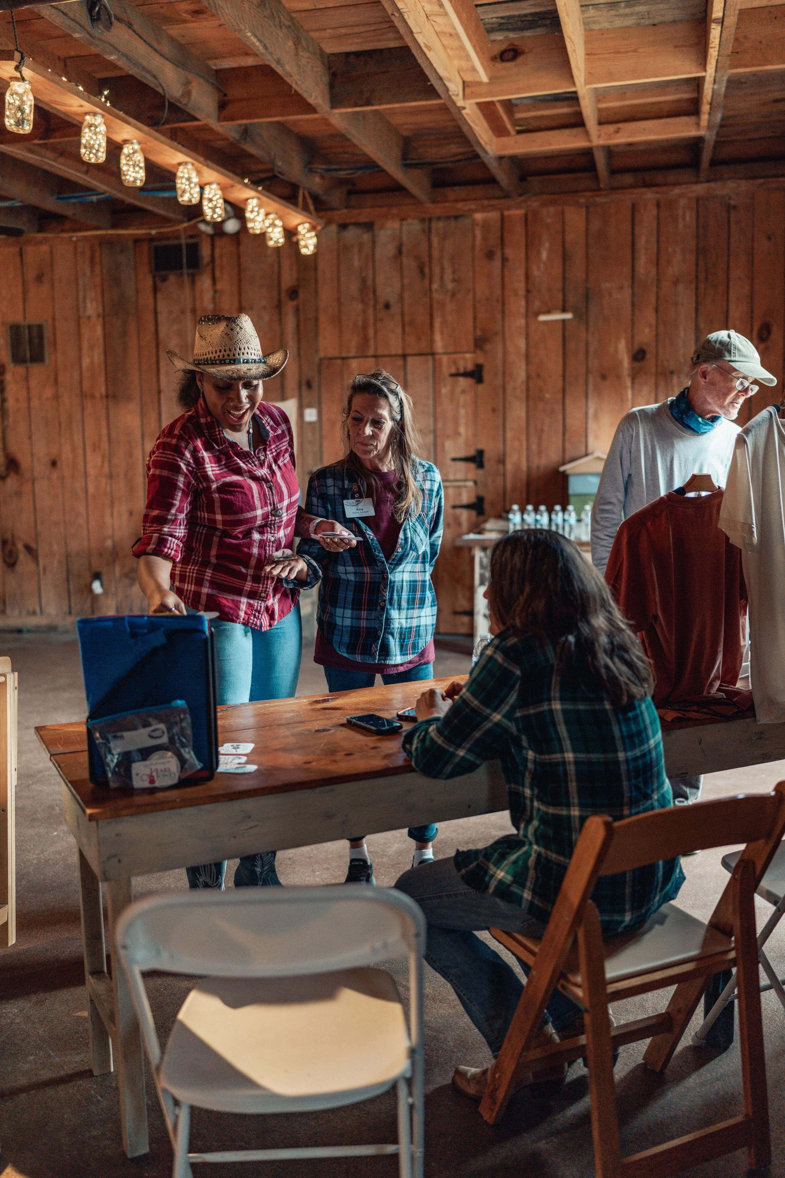 People gathered around a wooden table in a rustic, barn-like setting with wood-paneled walls and string lights hanging from the ceiling. Some people are standing and chatting, while others are seated.