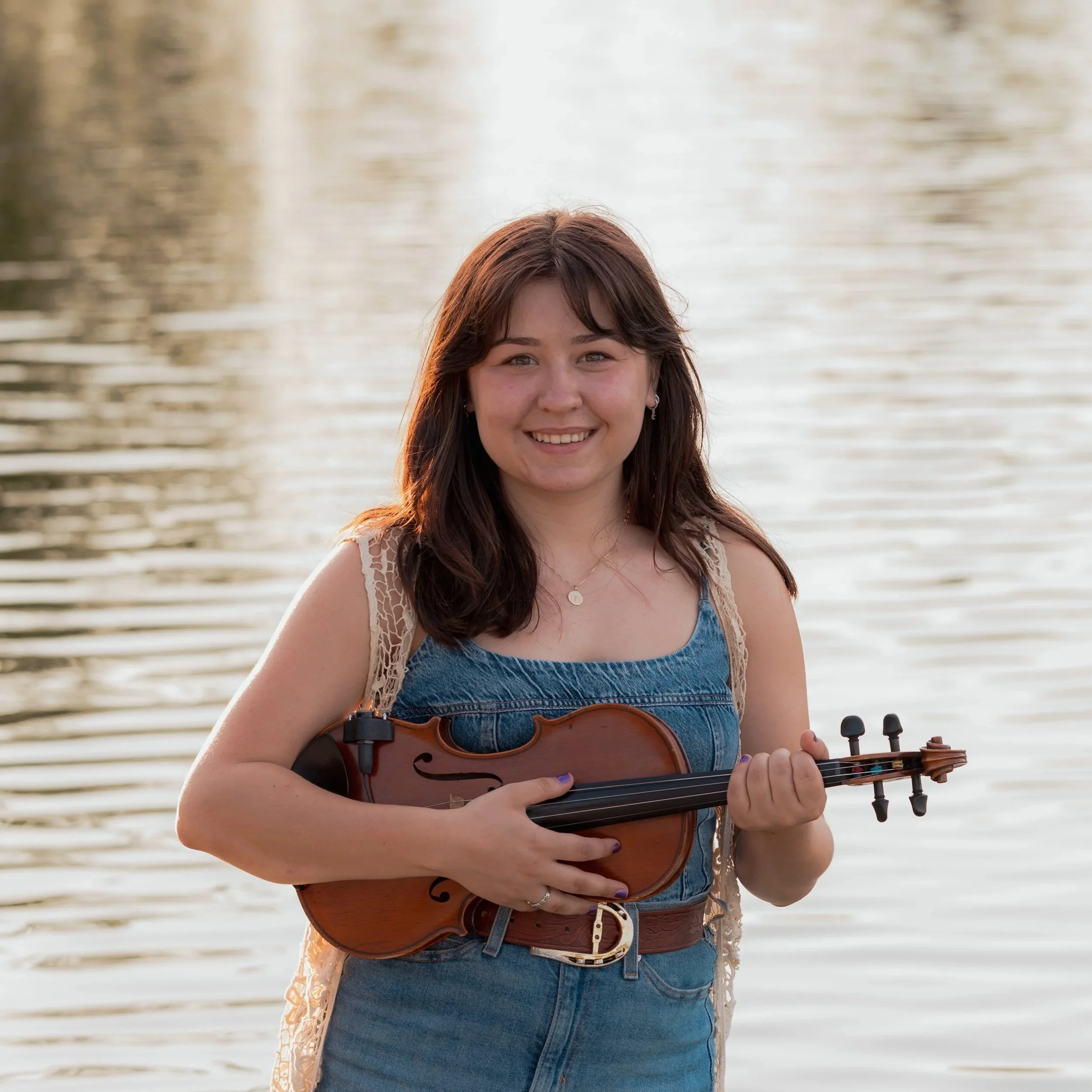 Young woman, Isabella,  with long brown hair holding a violin outdoors in sunlight, wearing a white lace dress, with a blurred natural background.