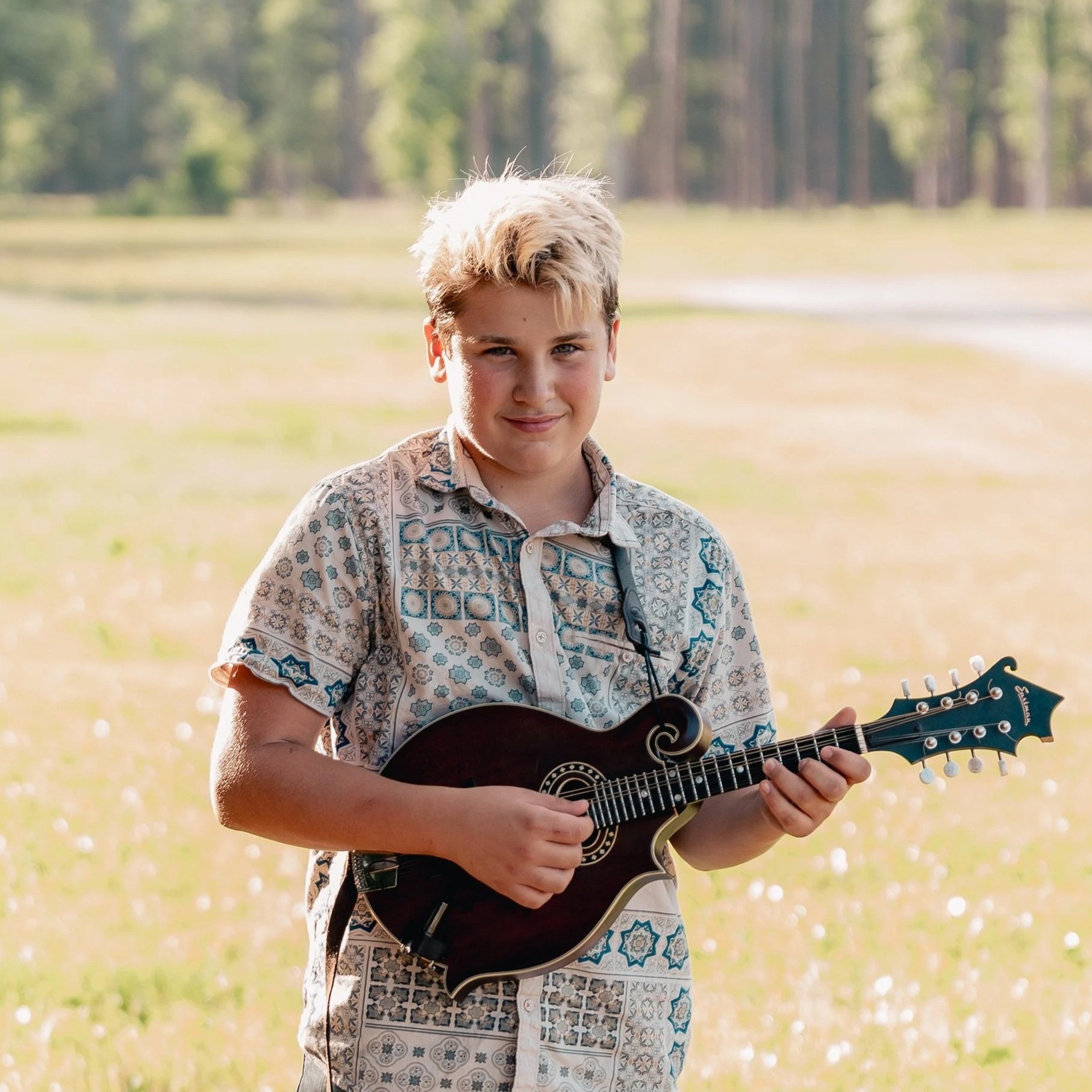 A young boy, Chris, with short blonde hair and a plaid shirt, sitting outdoors at sunset, playing a small mandolin.