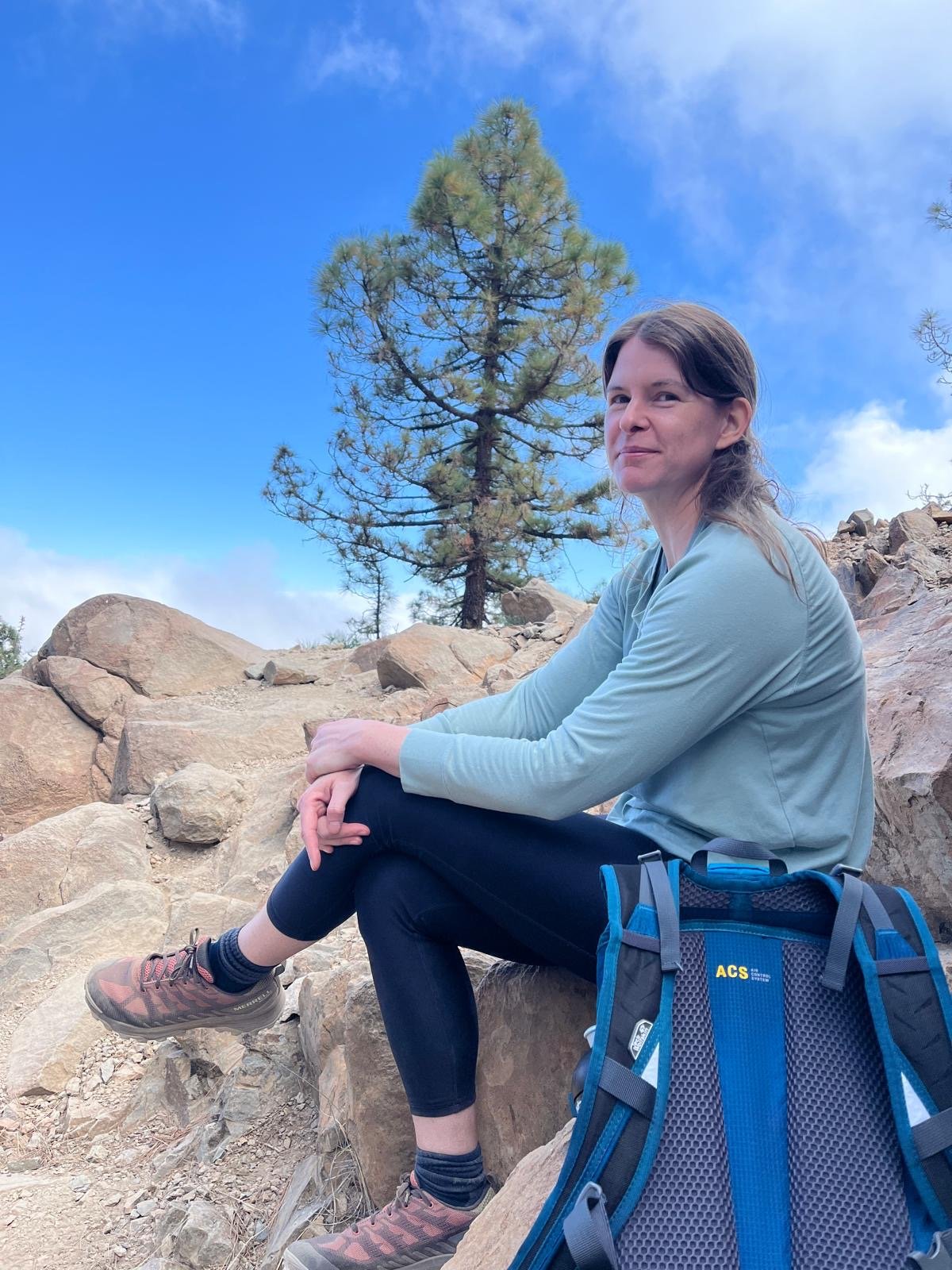 Een vrouw zit op rotsen tijdens een hike in de natuur, met een boom en een blauwe hemel op de achtergrond.