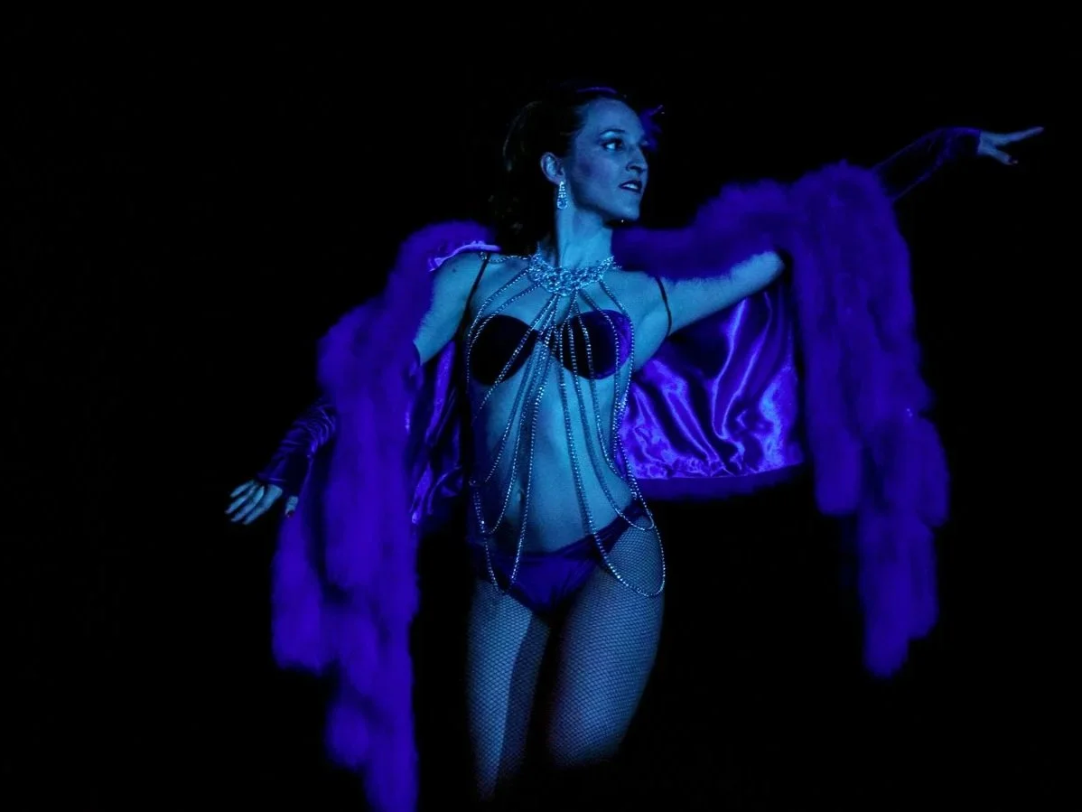 A woman performing on stage, dressed in purple lingerie, fishnet stockings, and a purple fur coat, with jewelry and dramatic makeup, under dark blue lighting.
