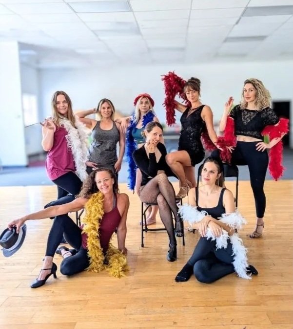 A group of seven women dressed in fashionable and colorful clothing, some wearing feather boas, posing in a spacious dance studio with a wooden floor.