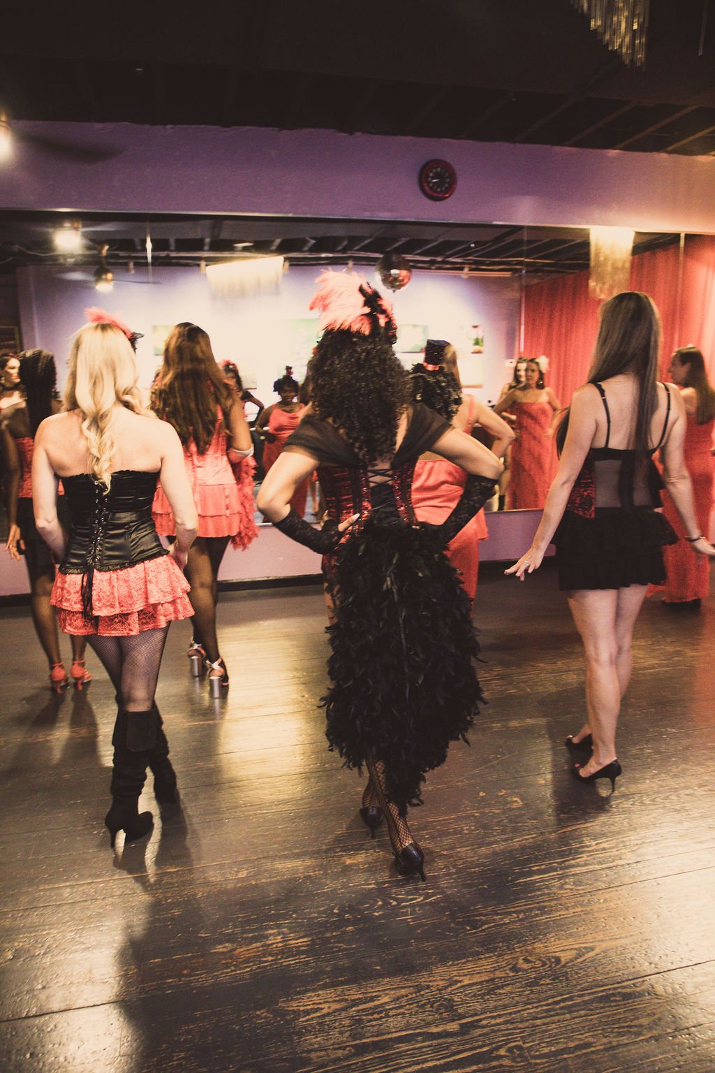 Women dressed in burlesque costumes dancing in a dimly lit room with a dance floor and control room in the background.