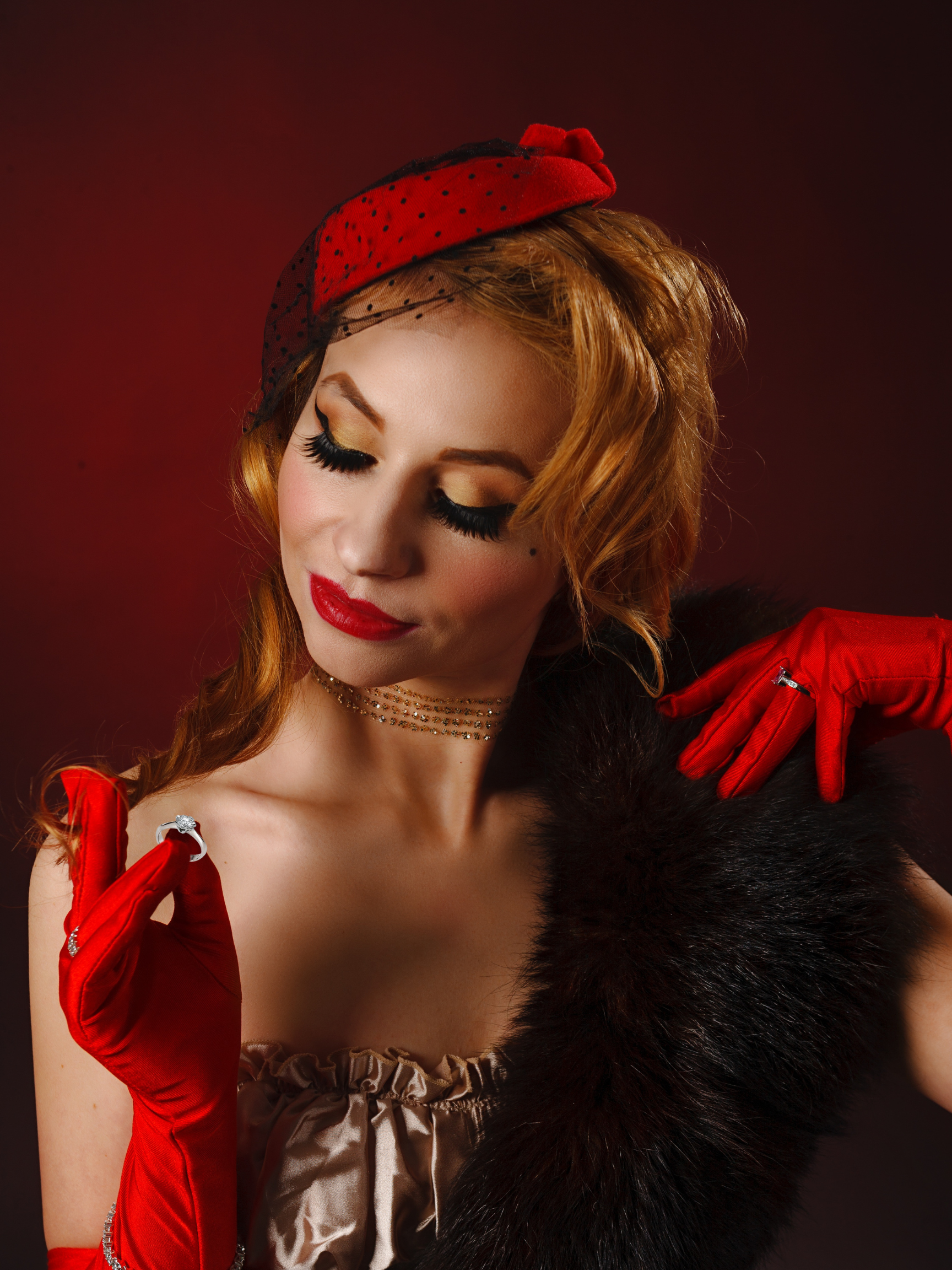 Portrait of a woman with red hair wearing red gloves, a fascinator, and a fur stole, with her eyes closed and a slight smile.