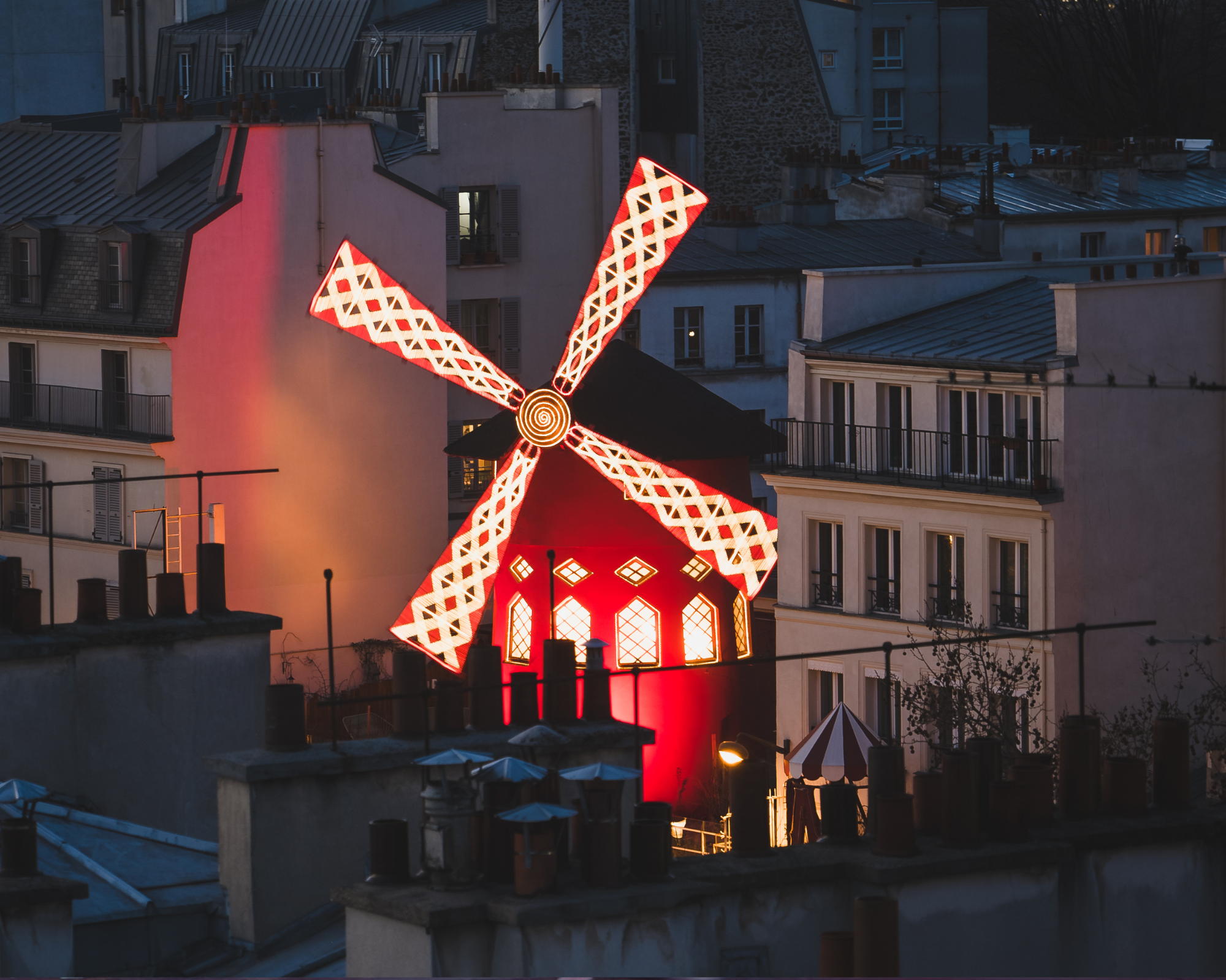 A lit-up decorative windmill with red and white patterns on its blades, mounted on a building rooftop in an urban area at dusk or night.