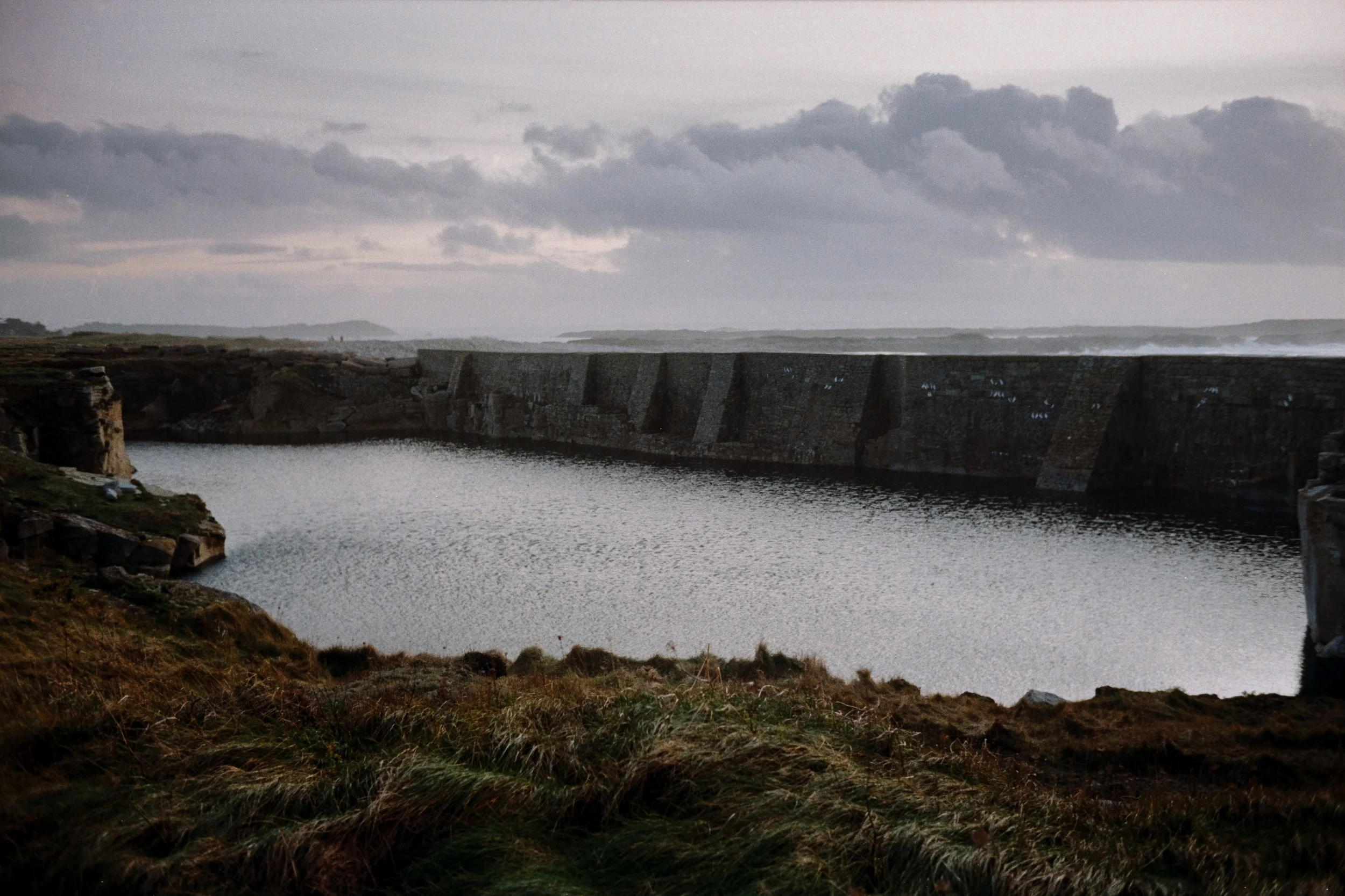 View of a stone dam in Brittany with water, grassy foreground, overcast sky.