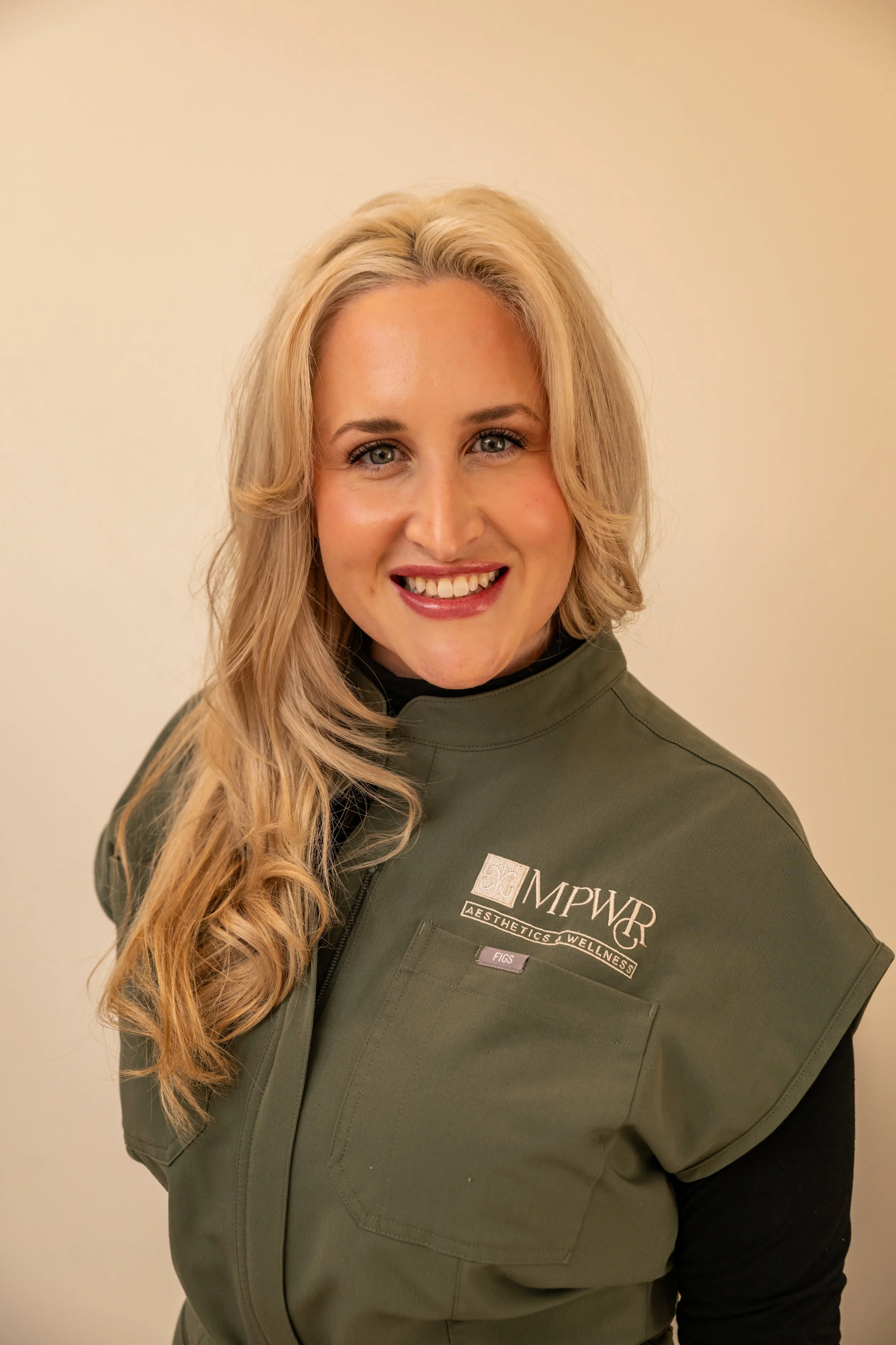 A woman with long blonde hair, blue eyes, and a friendly smile wearing a green vest with the logo 'MPWR Aesthetics & Wellness' and an ID badge that says 'FIGS'.