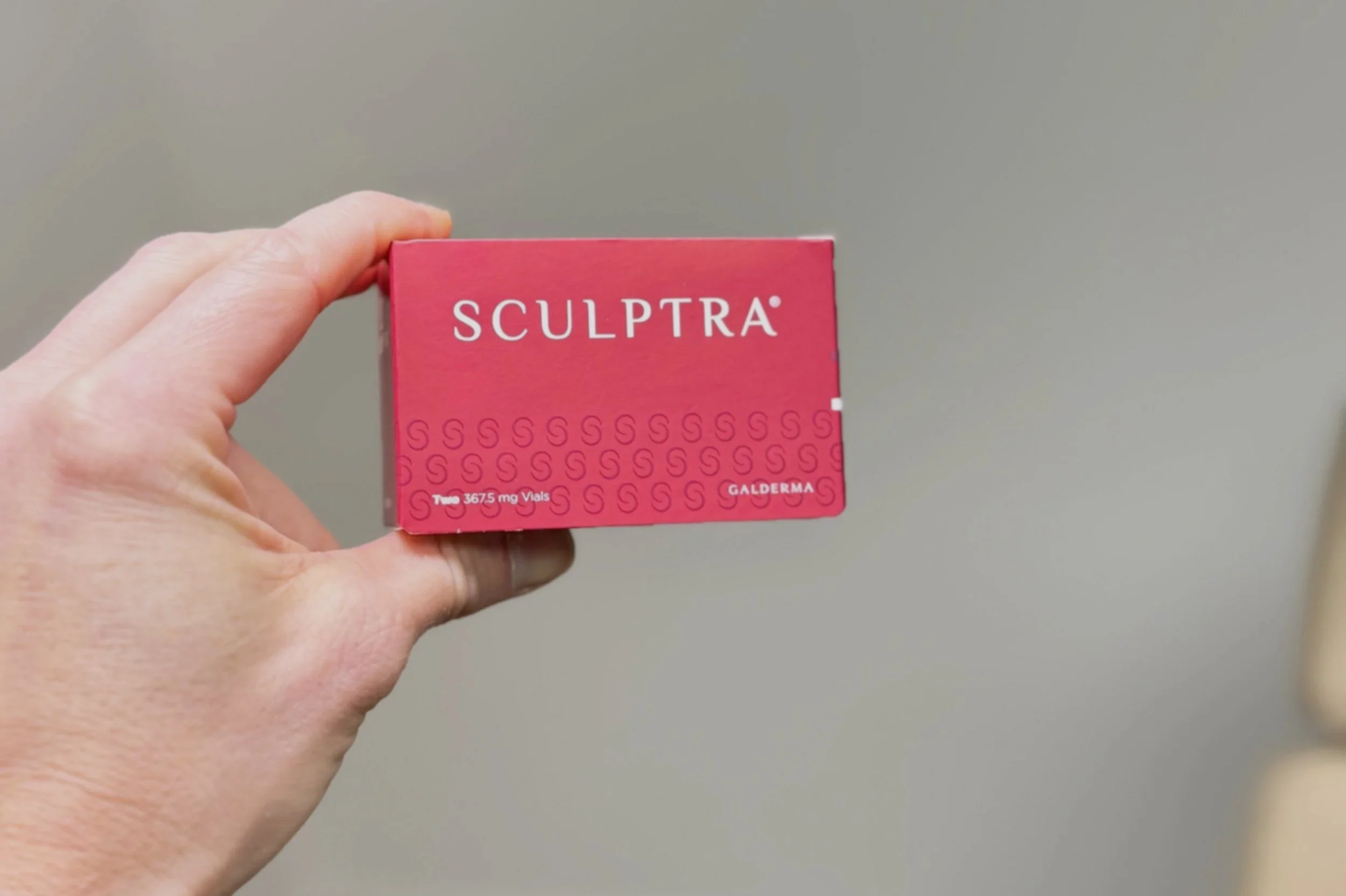 A person holding a pink box labeled 'Sculptra' against a plain background.