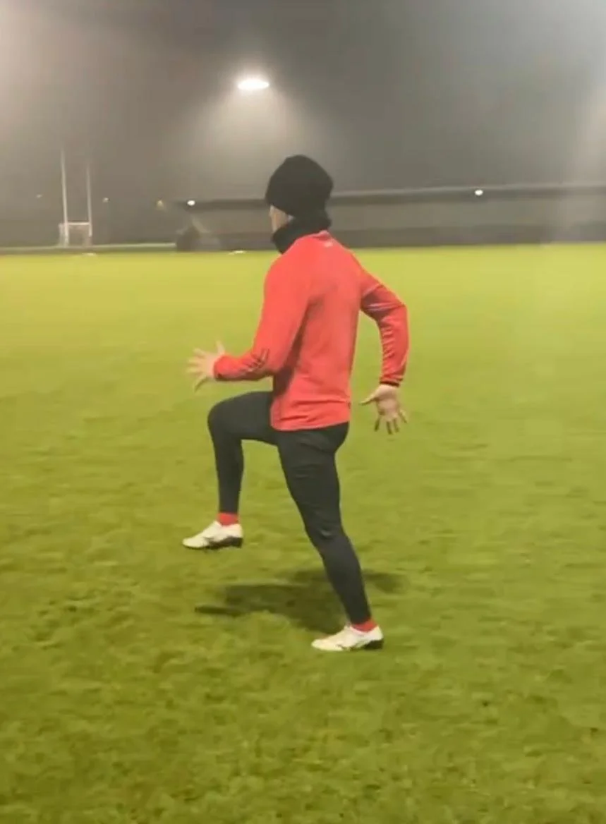 Person dressed in athletic clothing, including a red jacket, black pants, white shoes, a black beanie, and black gloves, performing a high knee exercise on a grass field at night.