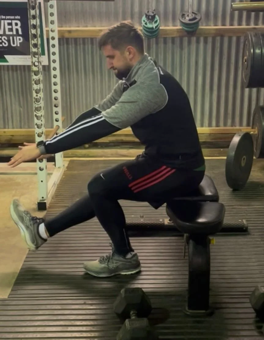 Man sitting on a kneeling exercise bench in a gym, stretching his leg forward while leaning slightly.