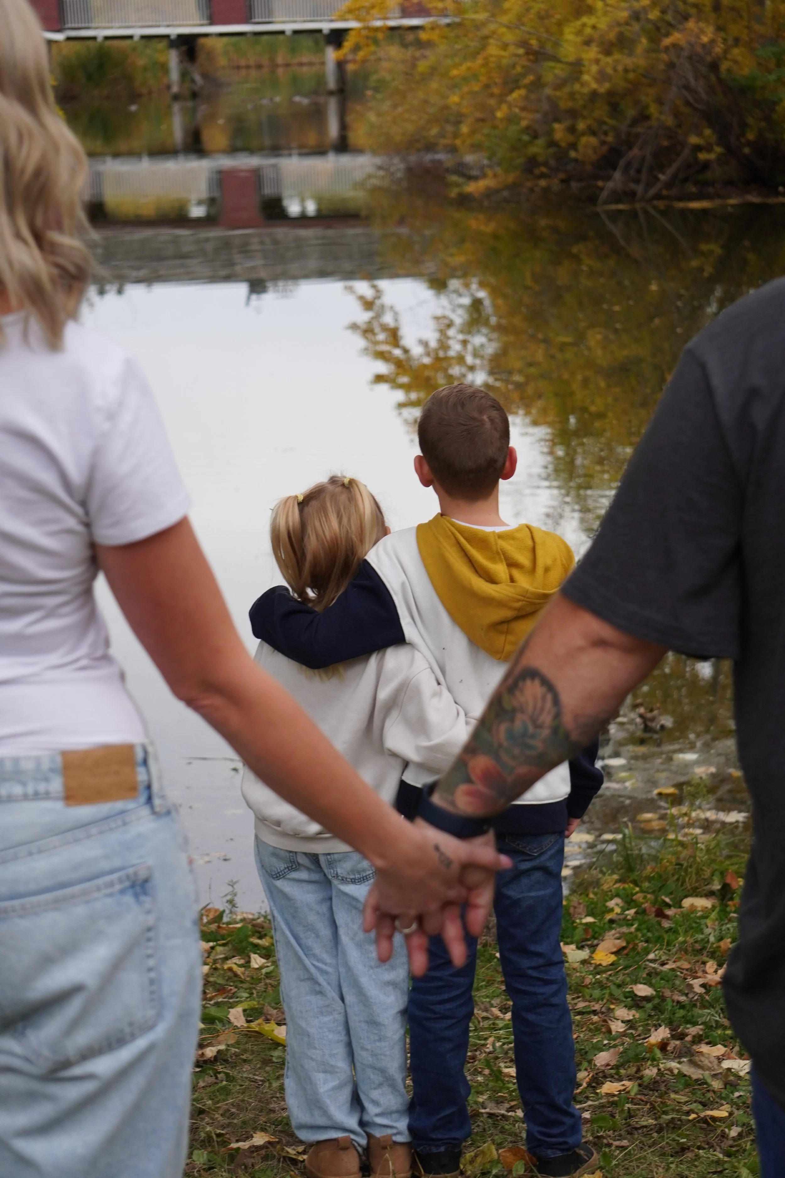 A family stands by a lake on an autumn day, holding hands and watching the water, with fall foliage in the background.