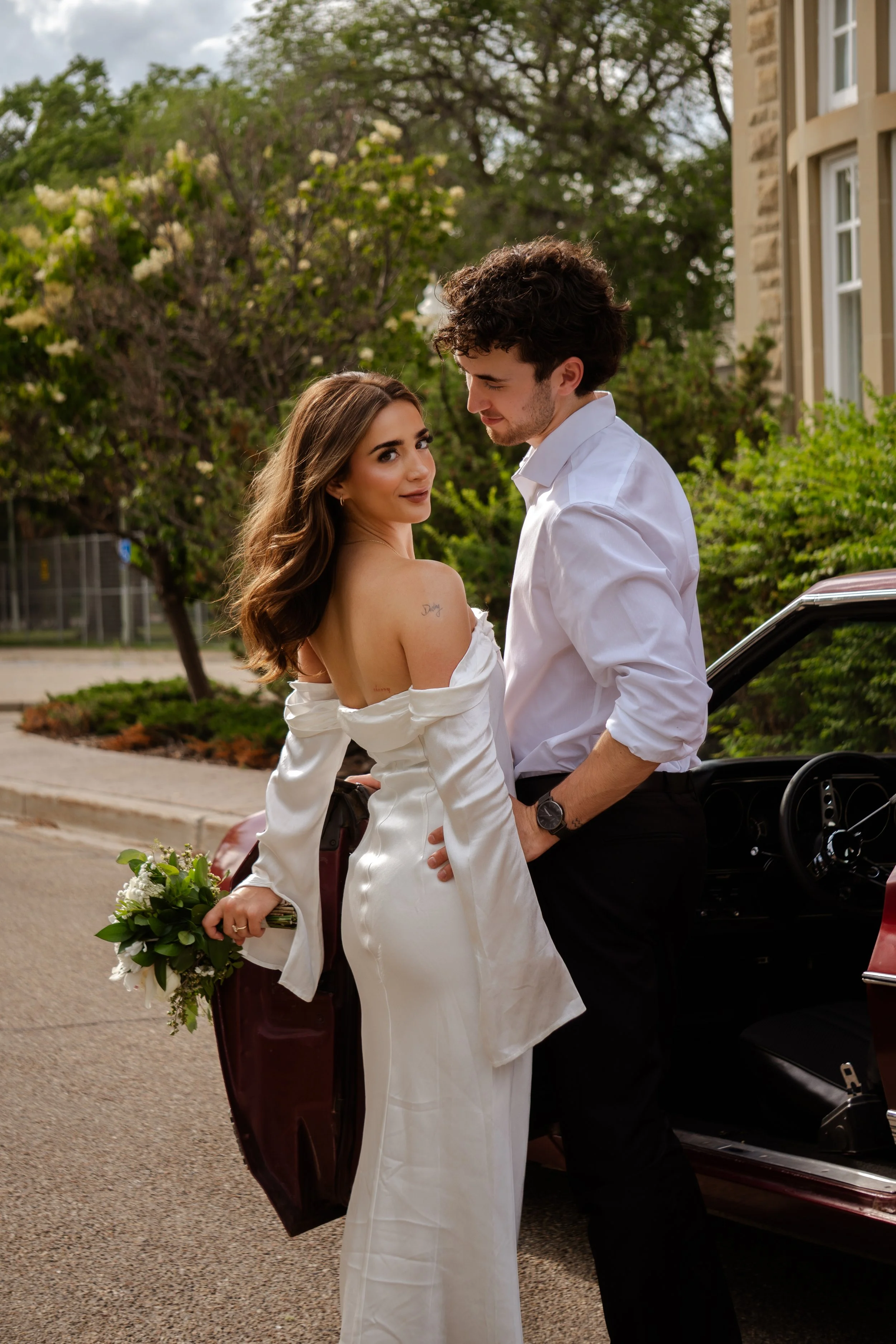 Edmonton wedding photography — bride with bouquet and groom beside vintage car outdoors.