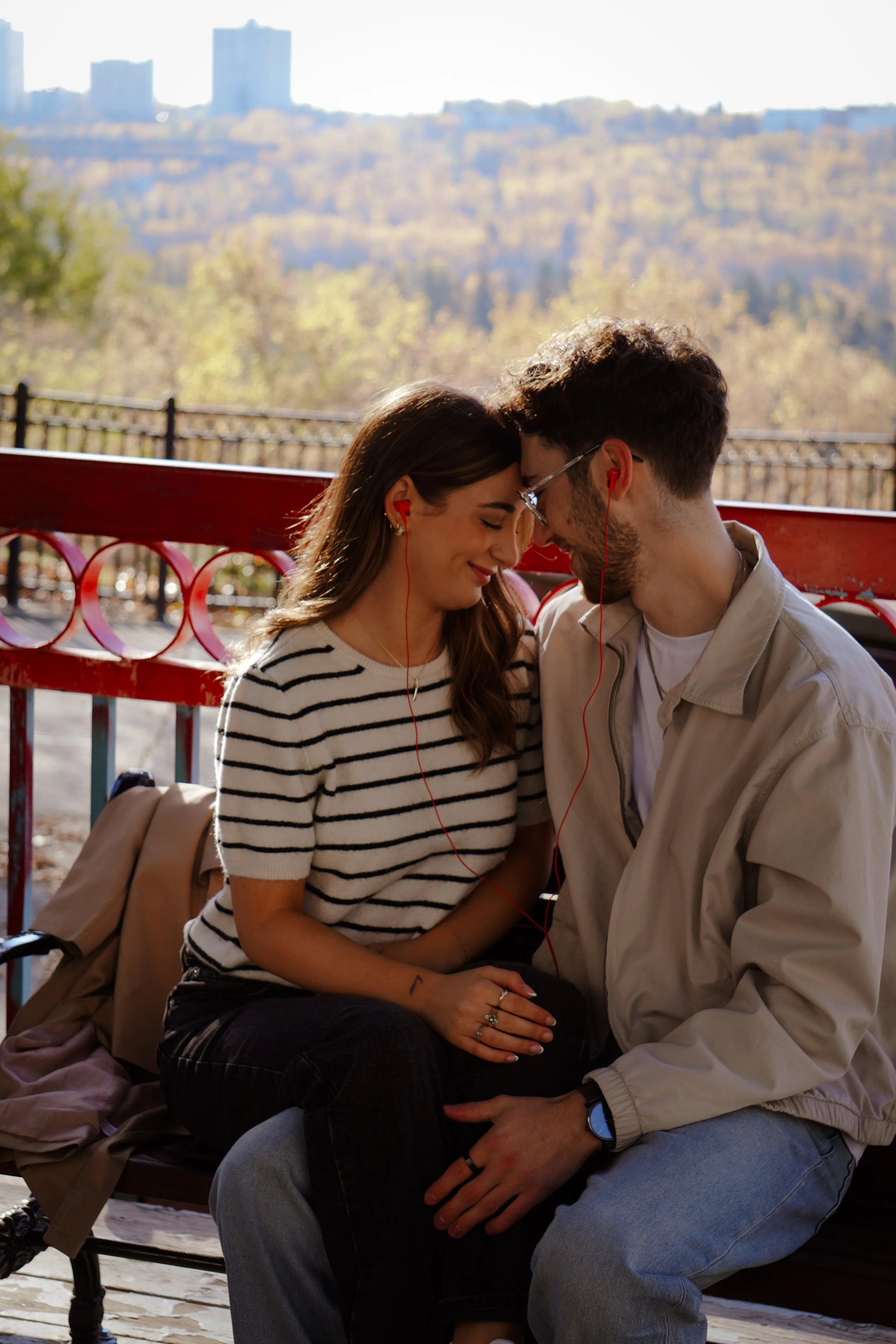 A young couple sitting close after listening to music with red earbuds, outdoors on a bench, with a city skyline and autumn trees in the background.