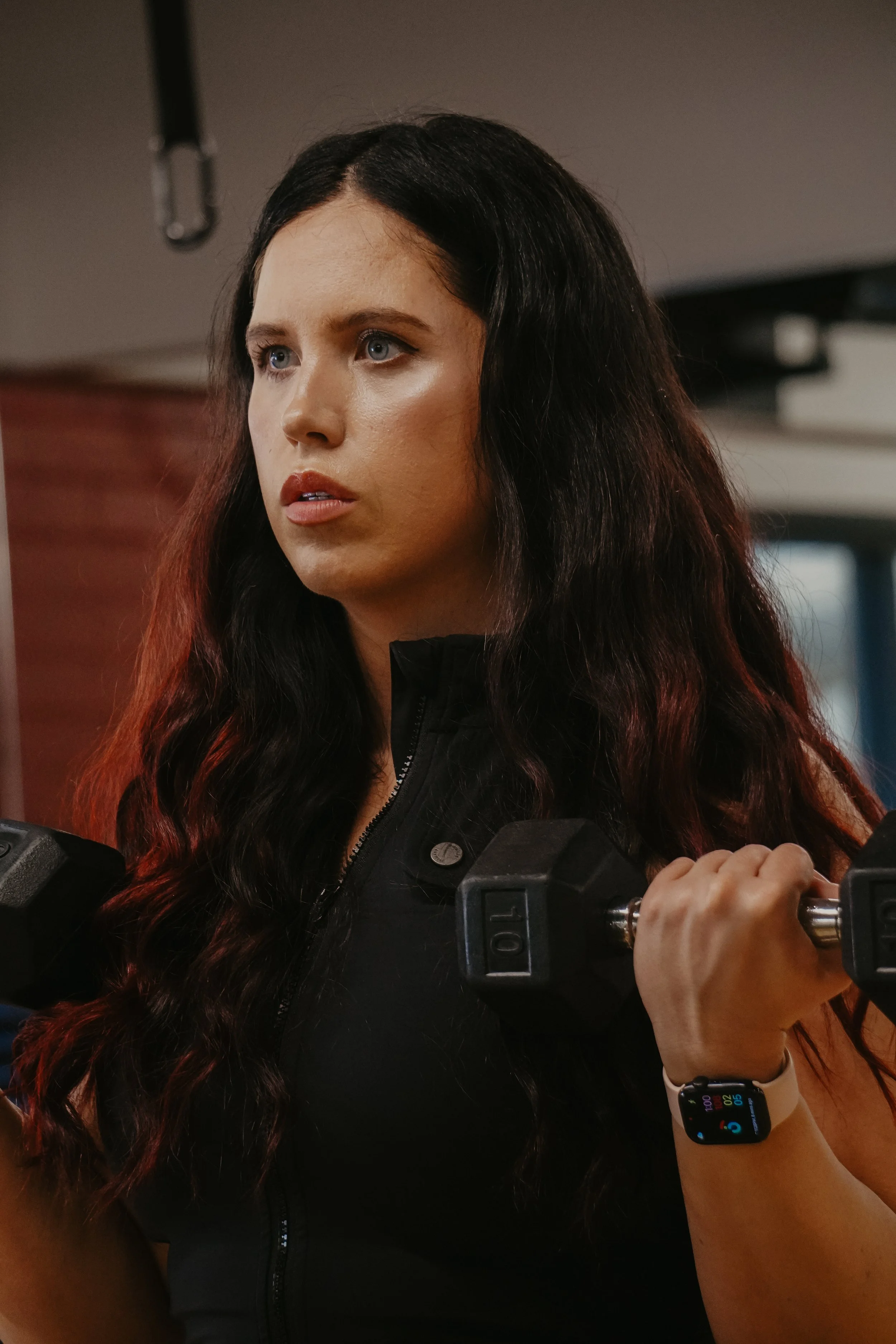 A woman with long dark wavy hair, wearing a black zip-up jacket and a smartwatch, holding a dumbbell in her right hand during workout.