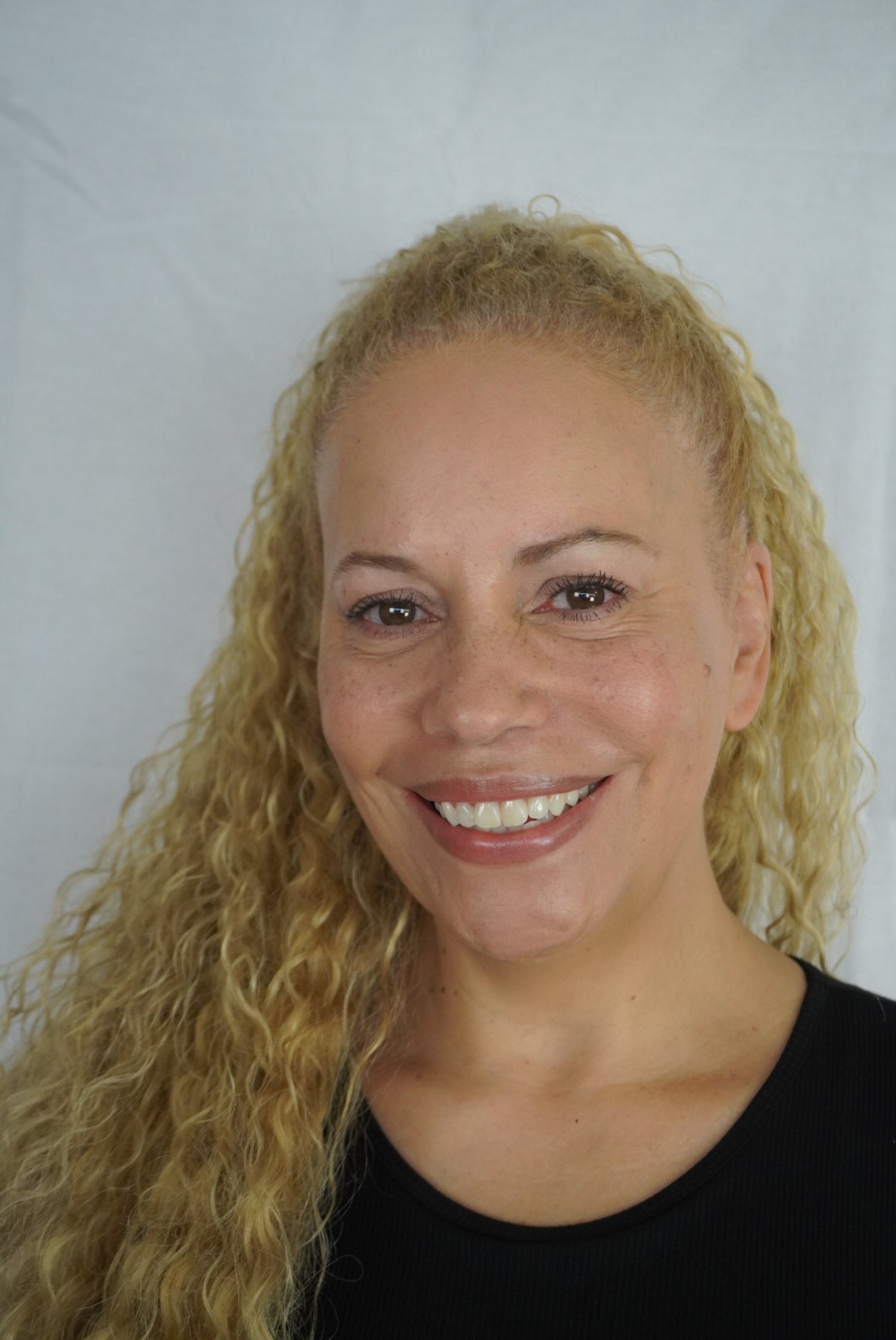 A woman with curly blonde hair smiling, wearing a black shirt, against a plain light-colored background.