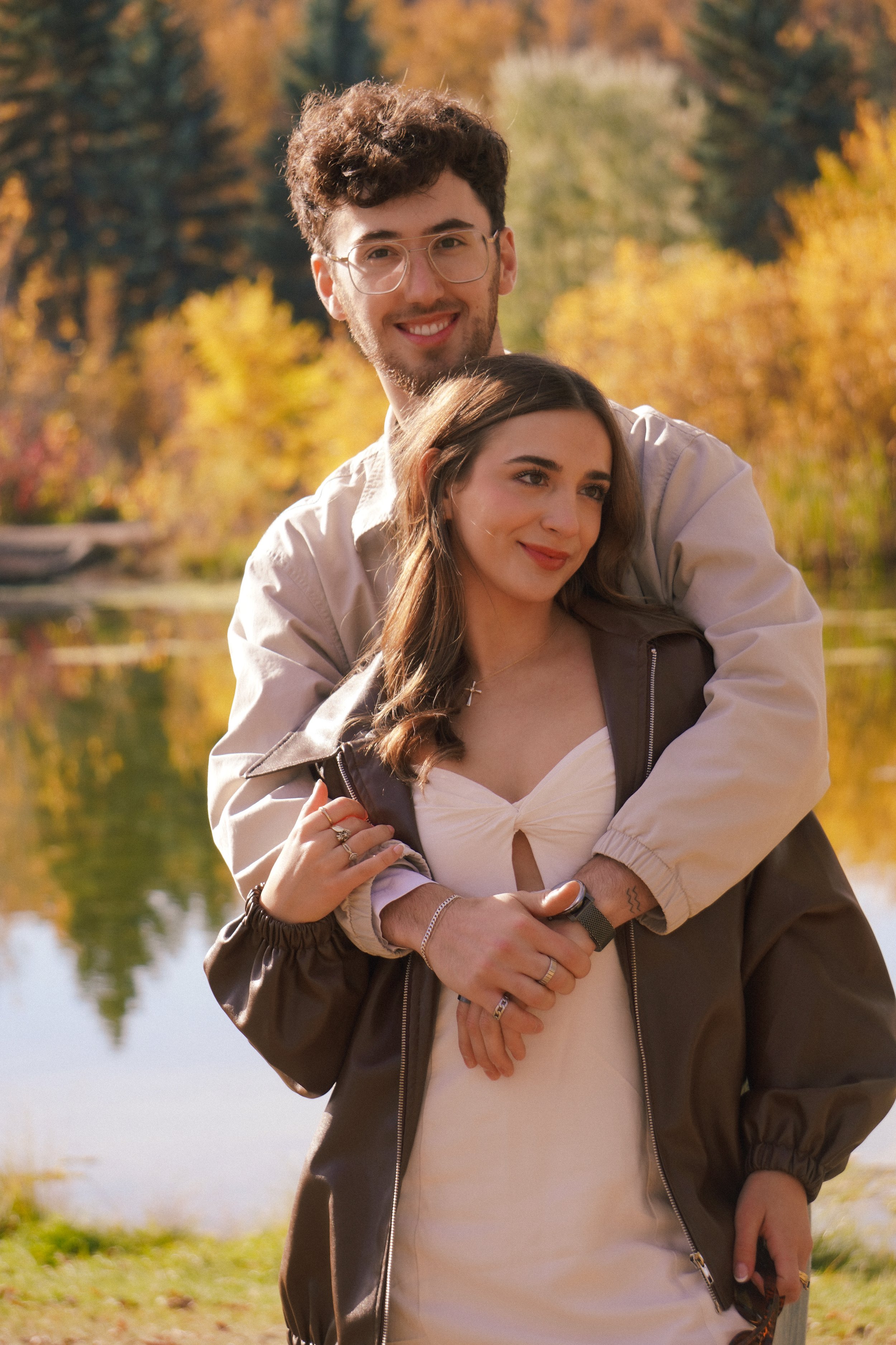 A young couple smiling and embracing outdoors near a lake with autumn trees in the background.