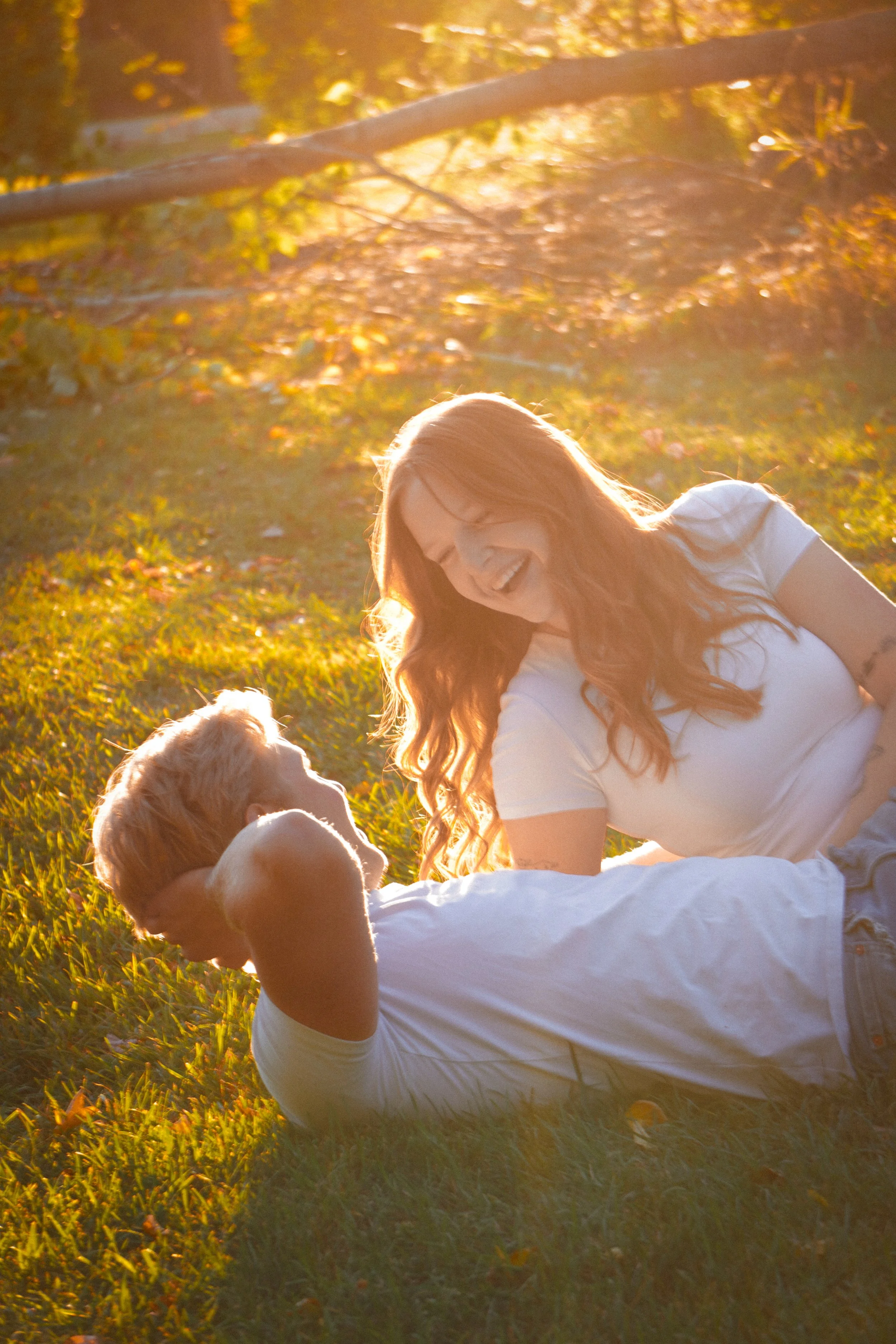 A woman and a man lying on the grass, smiling at each other in a park during sunset, with golden lighting and autumn leaves.