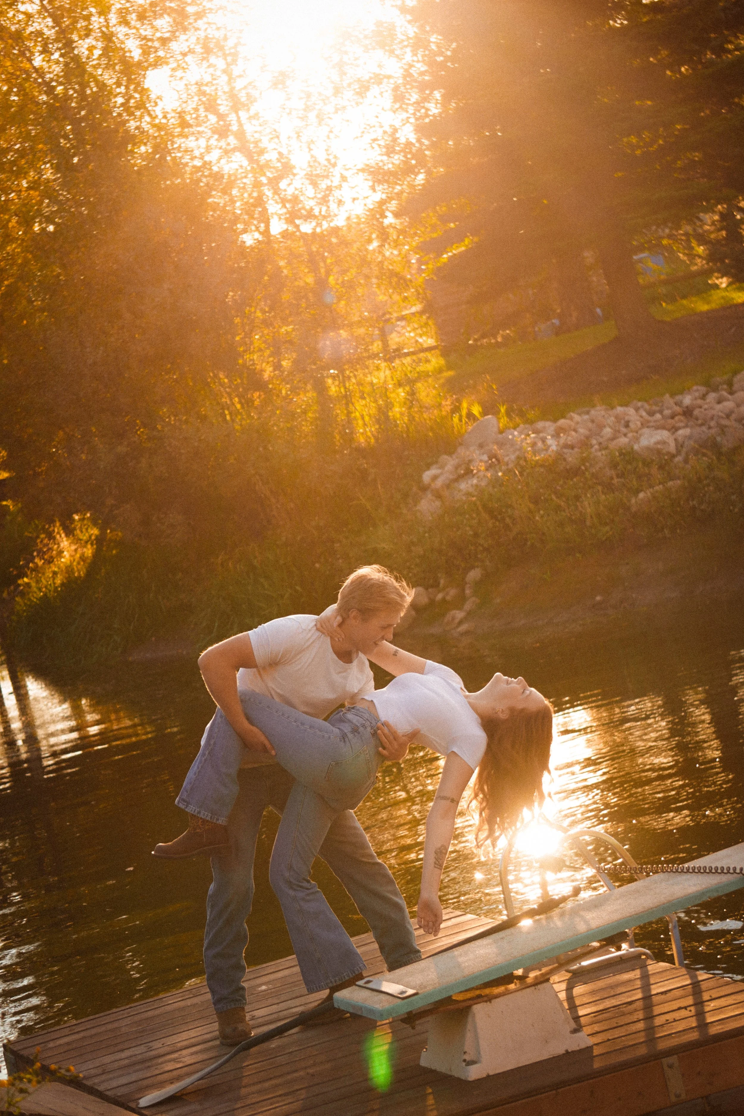 A young man is dipping a young woman backward on a dock by a lake during sunset, creating a romantic scene.