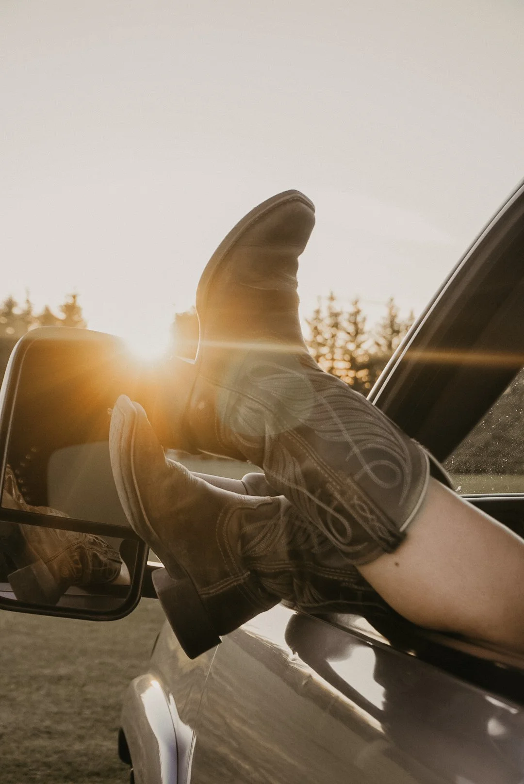 A person with cowboy boots resting their feet on the truck door window during sunset.