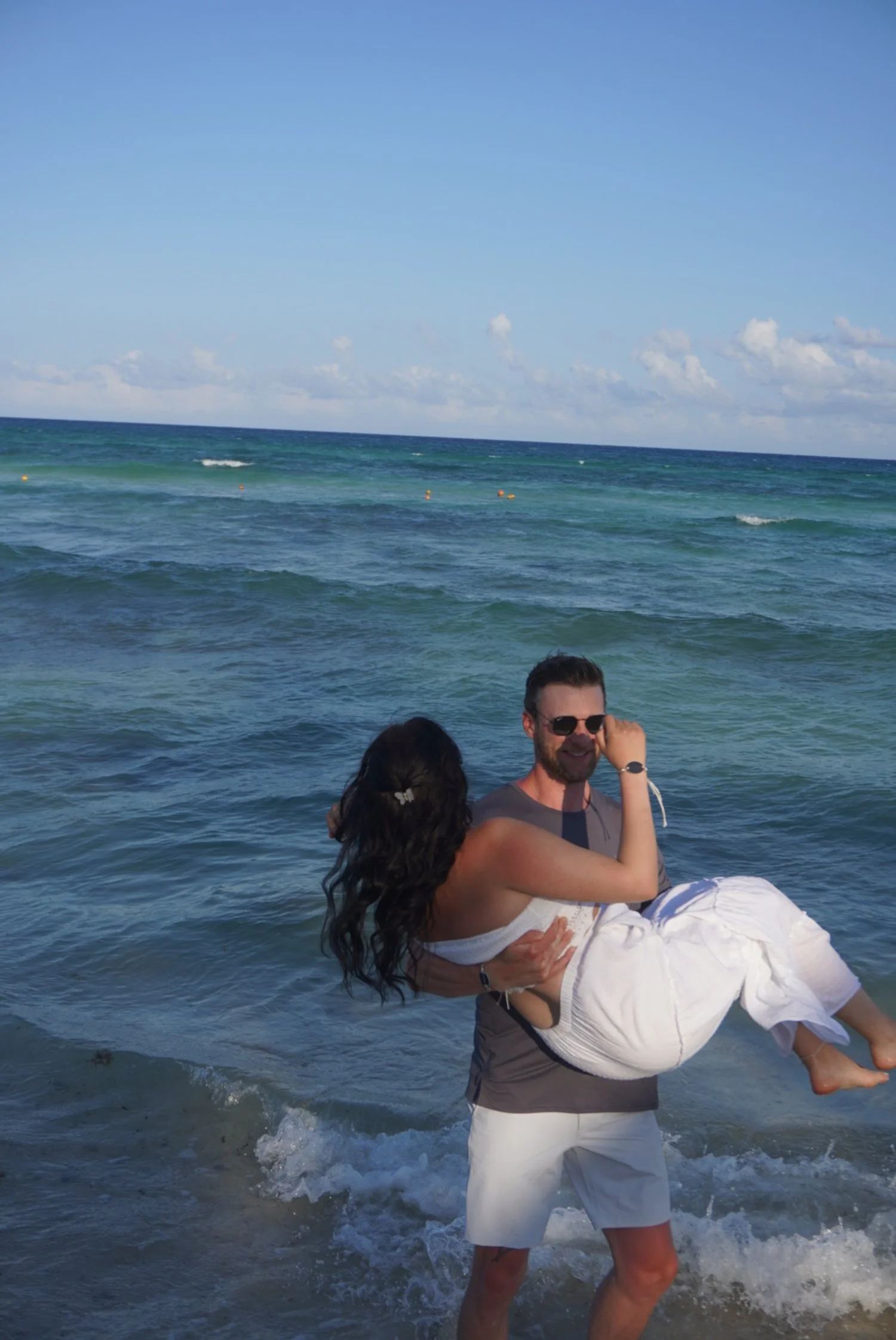 A man is holding a woman in his arms at the shoreline of a beach, with blue ocean waves and sky in the background.