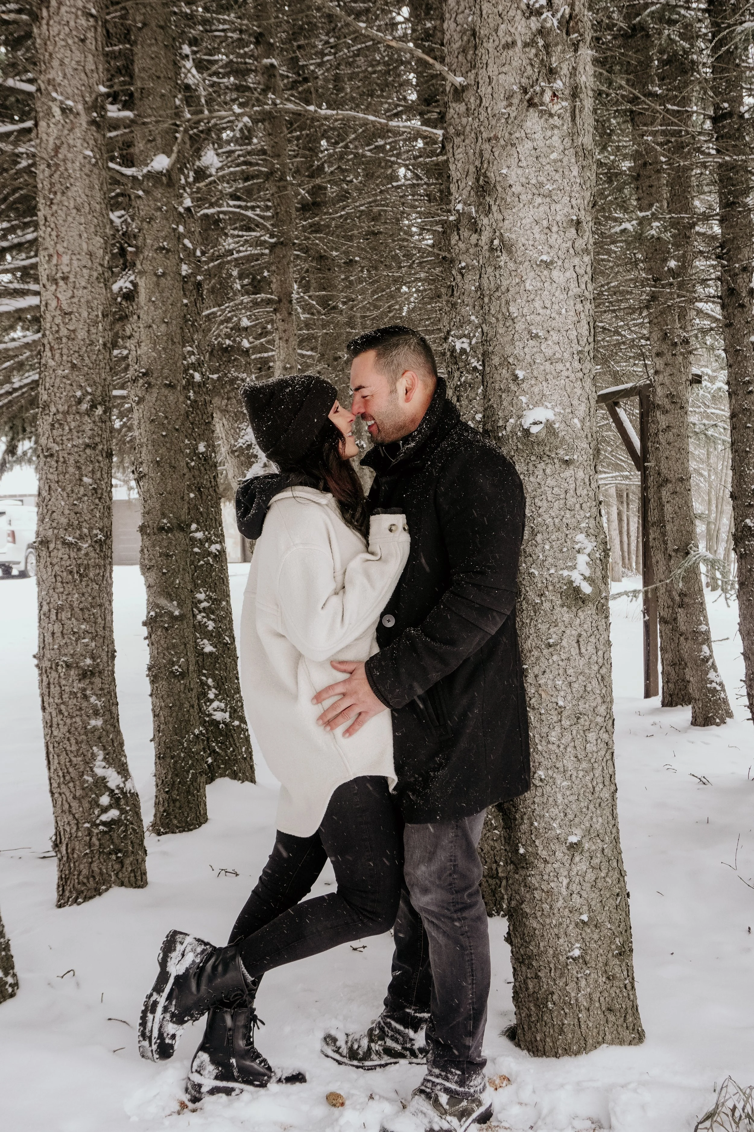 A couple laughing and leaning close to each other in a snowy forest, with trees and snow-covered ground in the background.