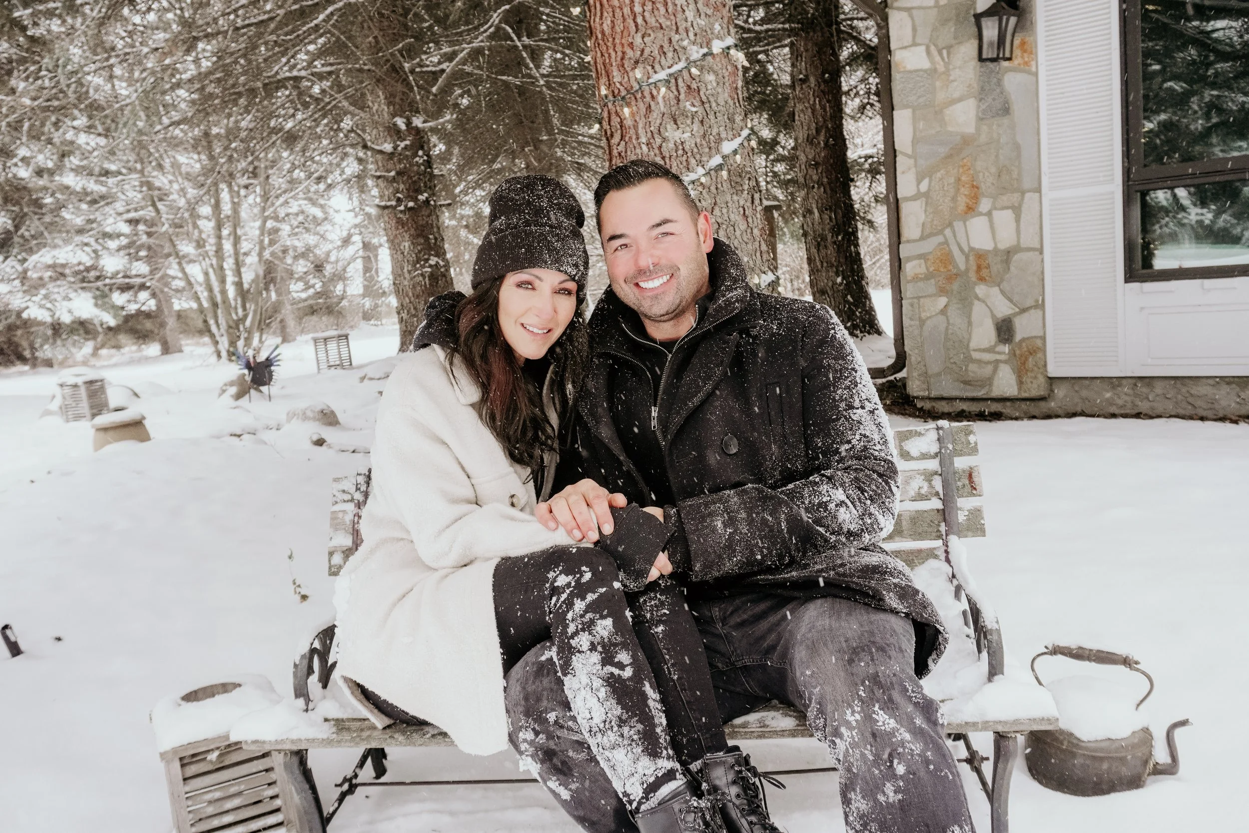 A smiling couple sitting on a wooden bench in a snowy outdoor setting, holding hands, with snow-covered trees and a stone house in the background.