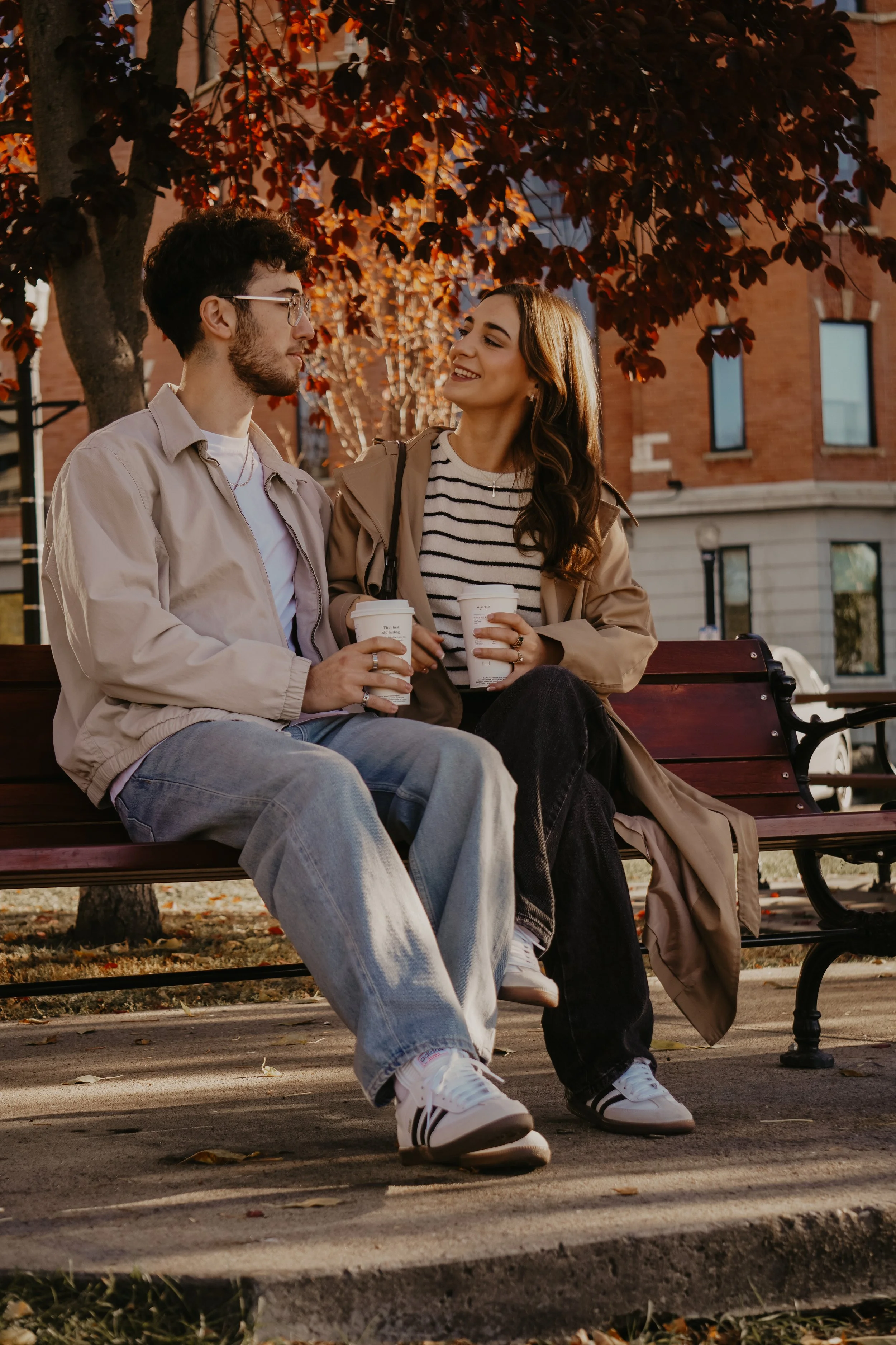 A young man and woman sit on a park bench, holding coffee cups, smiling at each other during autumn.