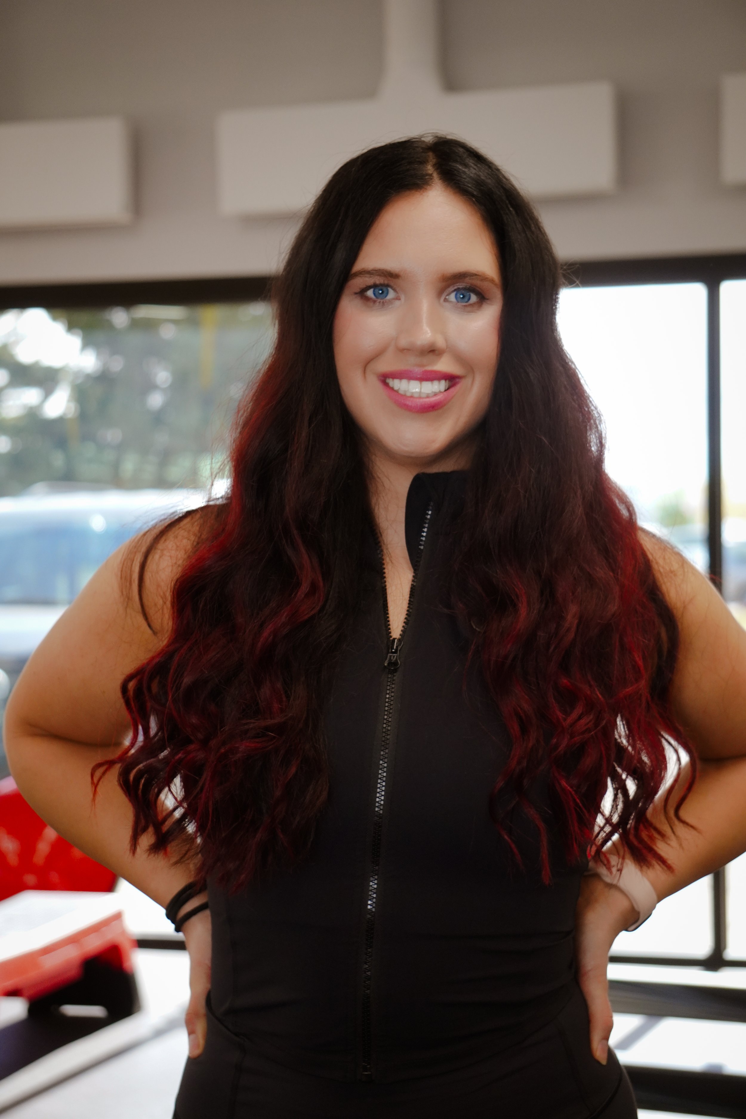 A woman with long, curly dark red hair with black roots, smiling and standing indoors near large windows.