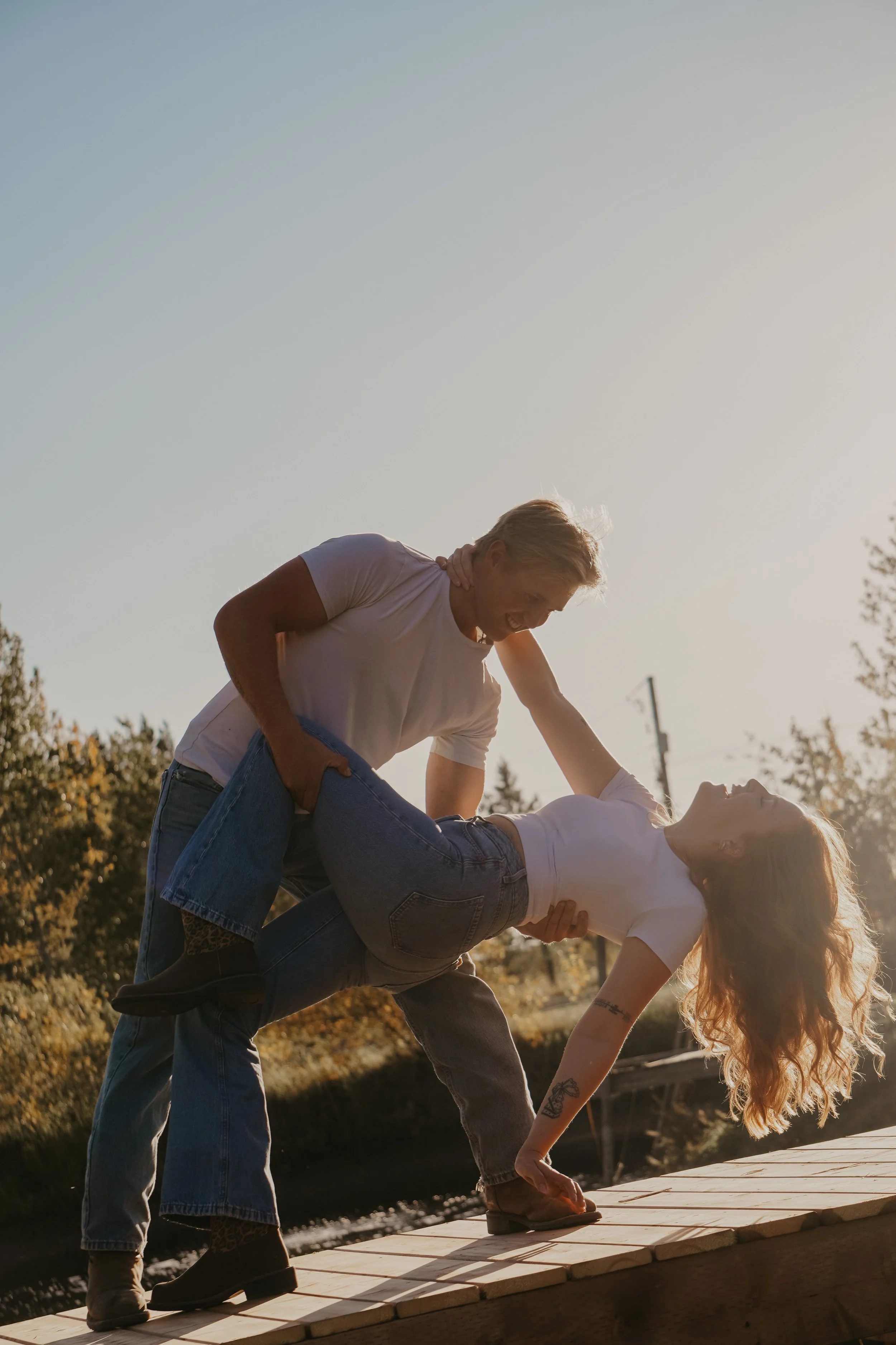 A man and woman engaged in a playful wrestling or dance scene outdoors during sunset, with trees and a body of water in the background.
