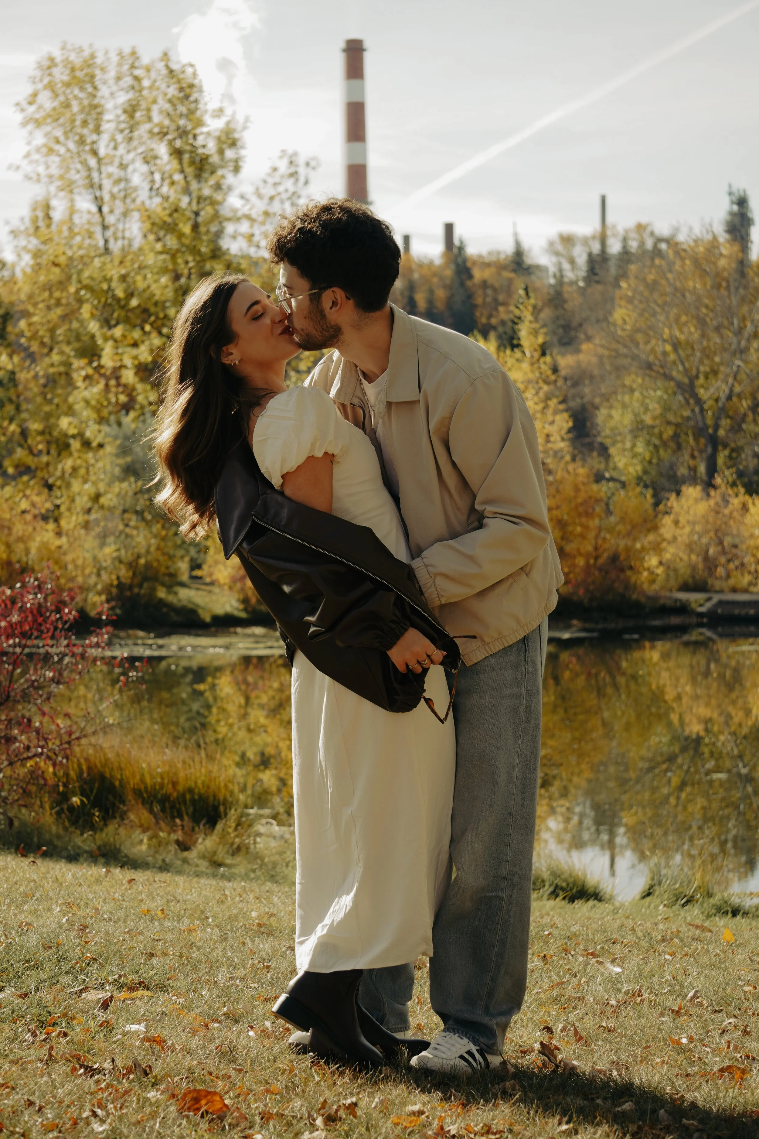 A couple sharing a kiss outdoors by a lake with autumn trees and a smokestack in the background.