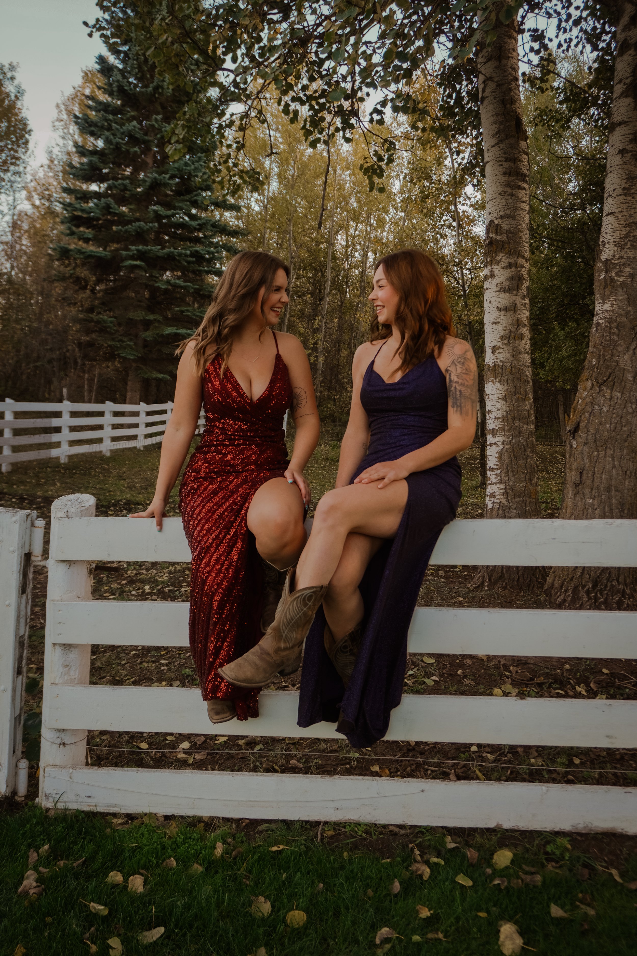 Two women in evening dresses sitting on a white wooden fence outdoors, smiling and talking, with trees and a white picket fence in the background.