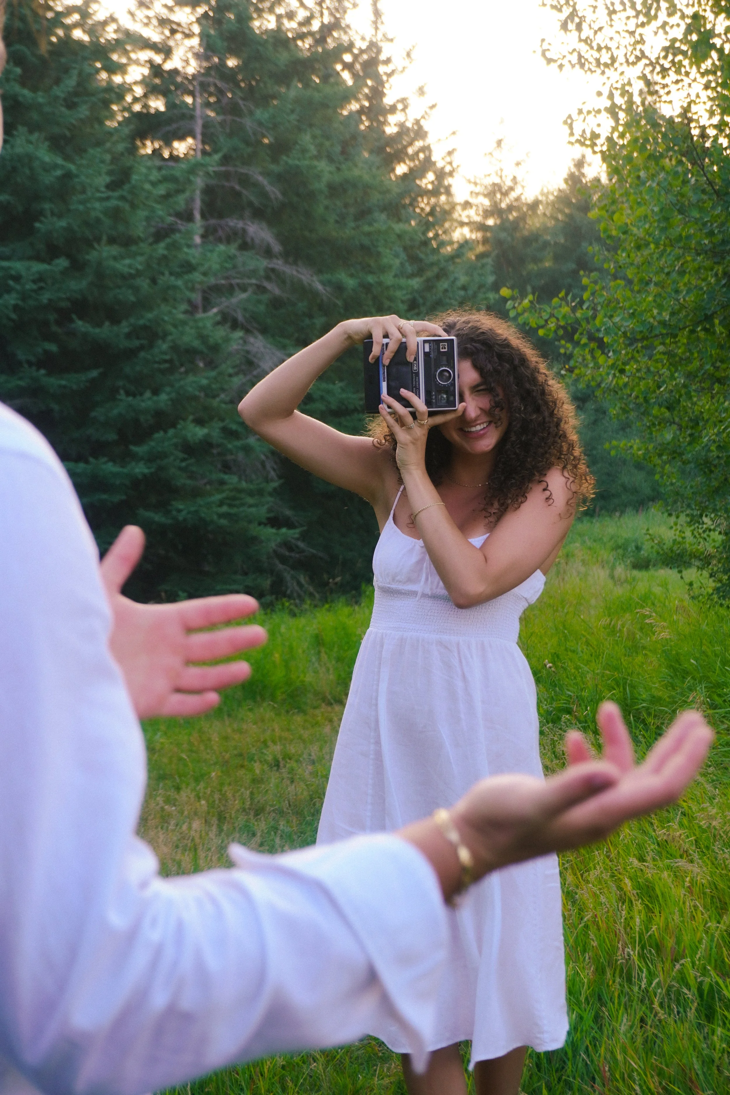 A woman with curly hair in a white dress is smiling and taking a photo with a vintage camera in a green outdoor setting, with trees and sunlight in the background.