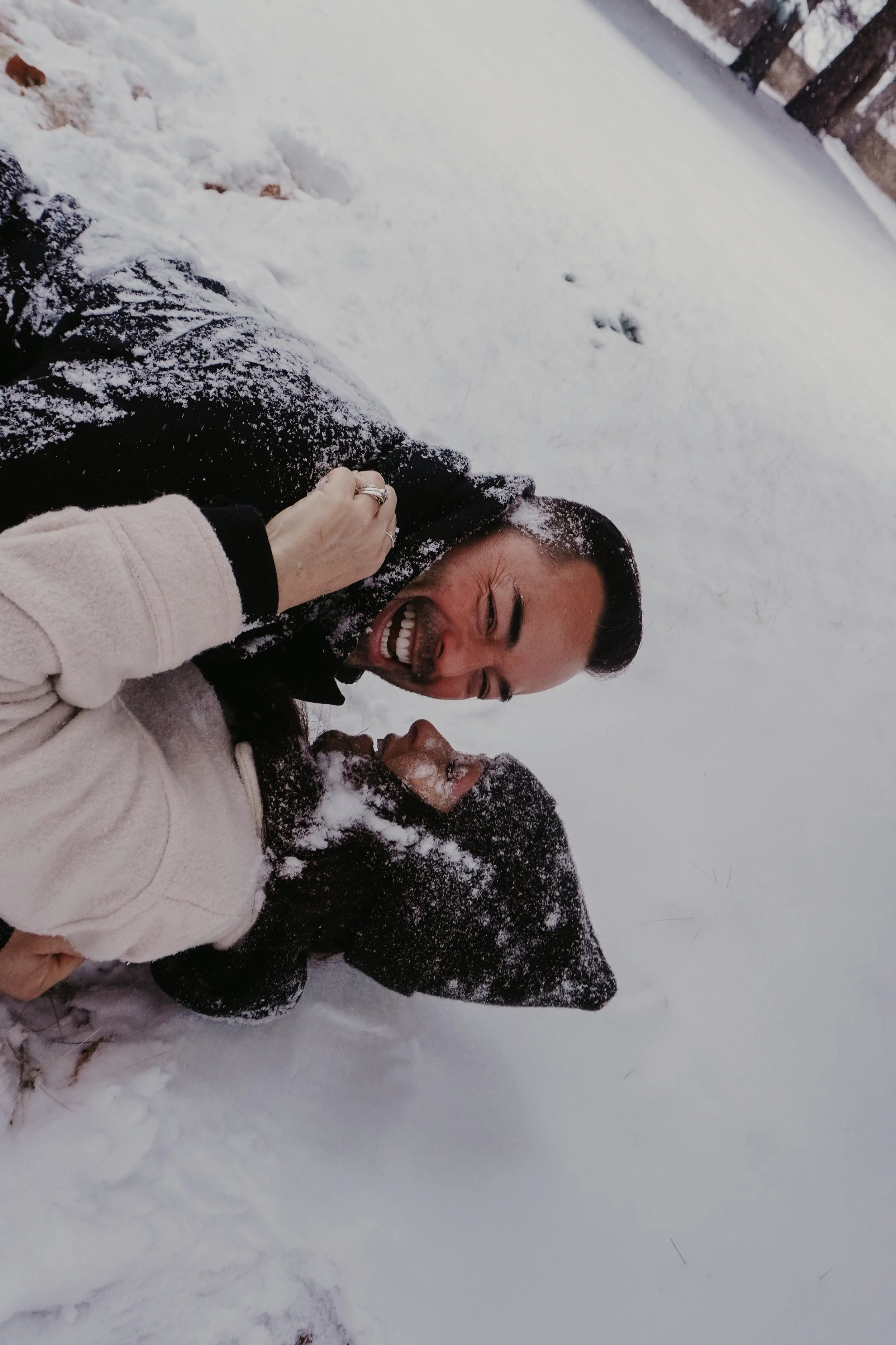 Two people in a snow-covered outdoor setting, smiling and playing in the snow, with snow on their faces and clothing.