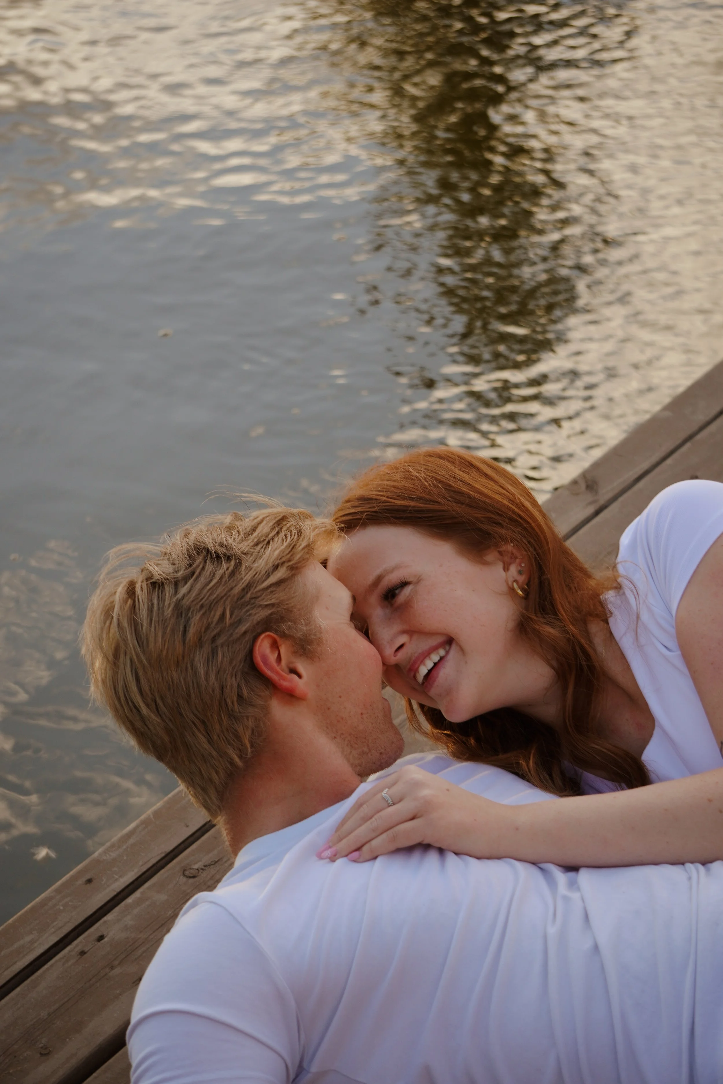 A couple lying on a dock by the water, smiling and touching foreheads, during sunset.