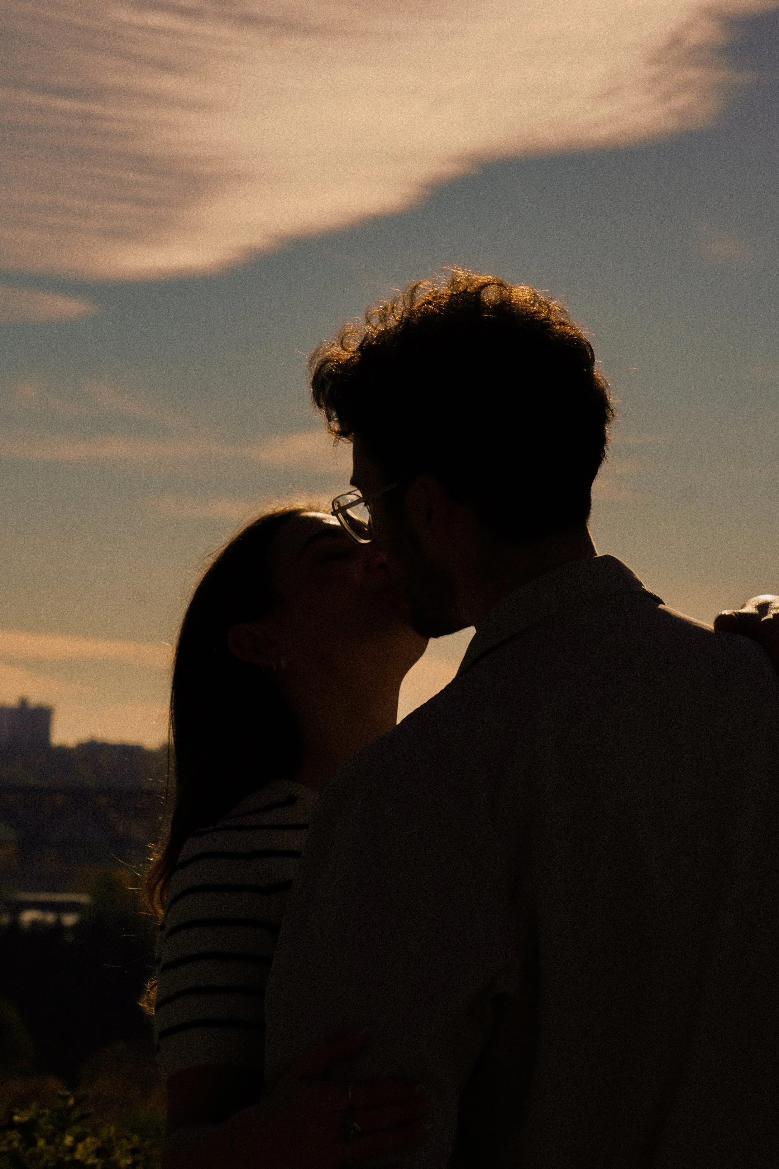 Silhouette of a couple kissing outdoors during sunset with clouds in the sky.