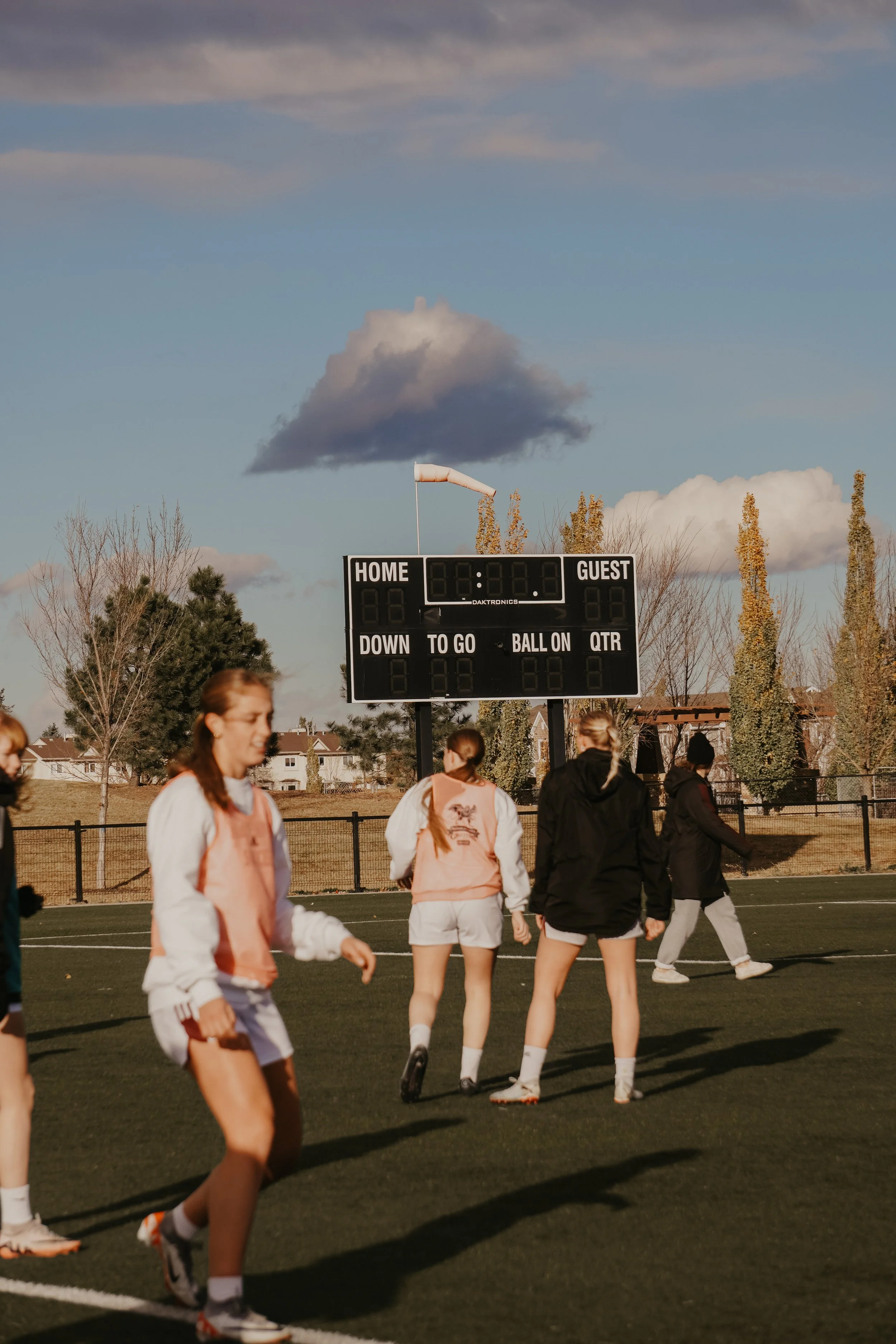 Girl soccer players warming up on a field with an electronic scoreboard in the background showing 00 for home and guest scores under a partly cloudy sky.
