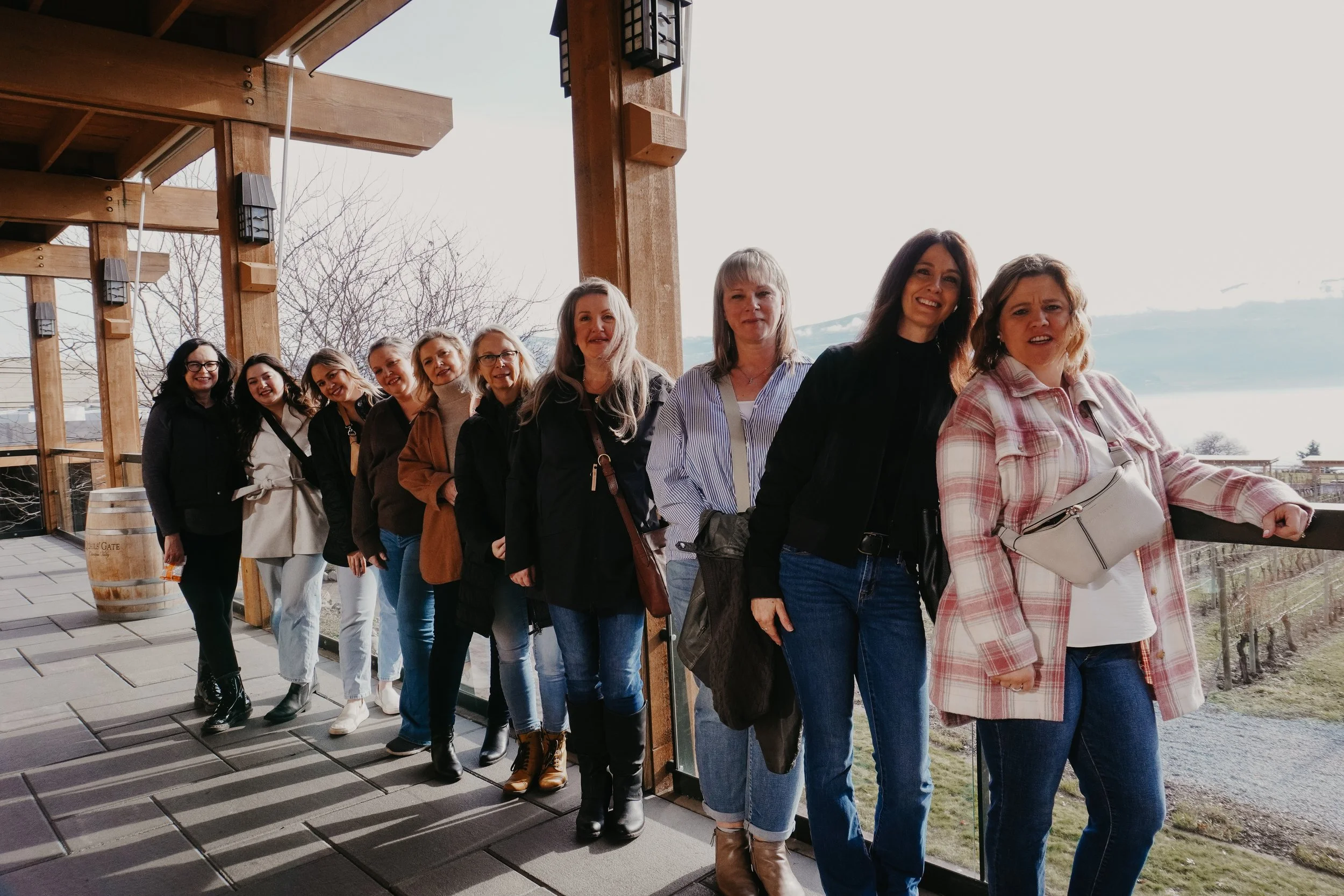 A group of eleven women standing in a line on a balcony with wooden beams, overlooking a vineyard and a body of water in the background.