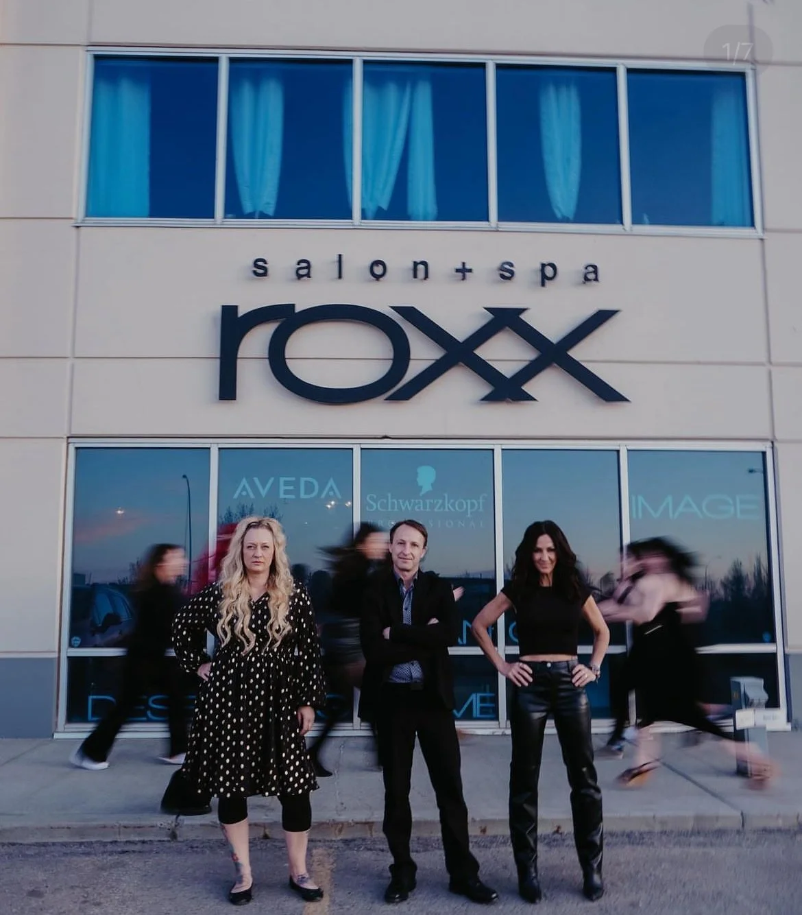 Three people stand in front of a building with sign 'salon+spa Roxy' above the windows, with blurred figures passing by in the background.