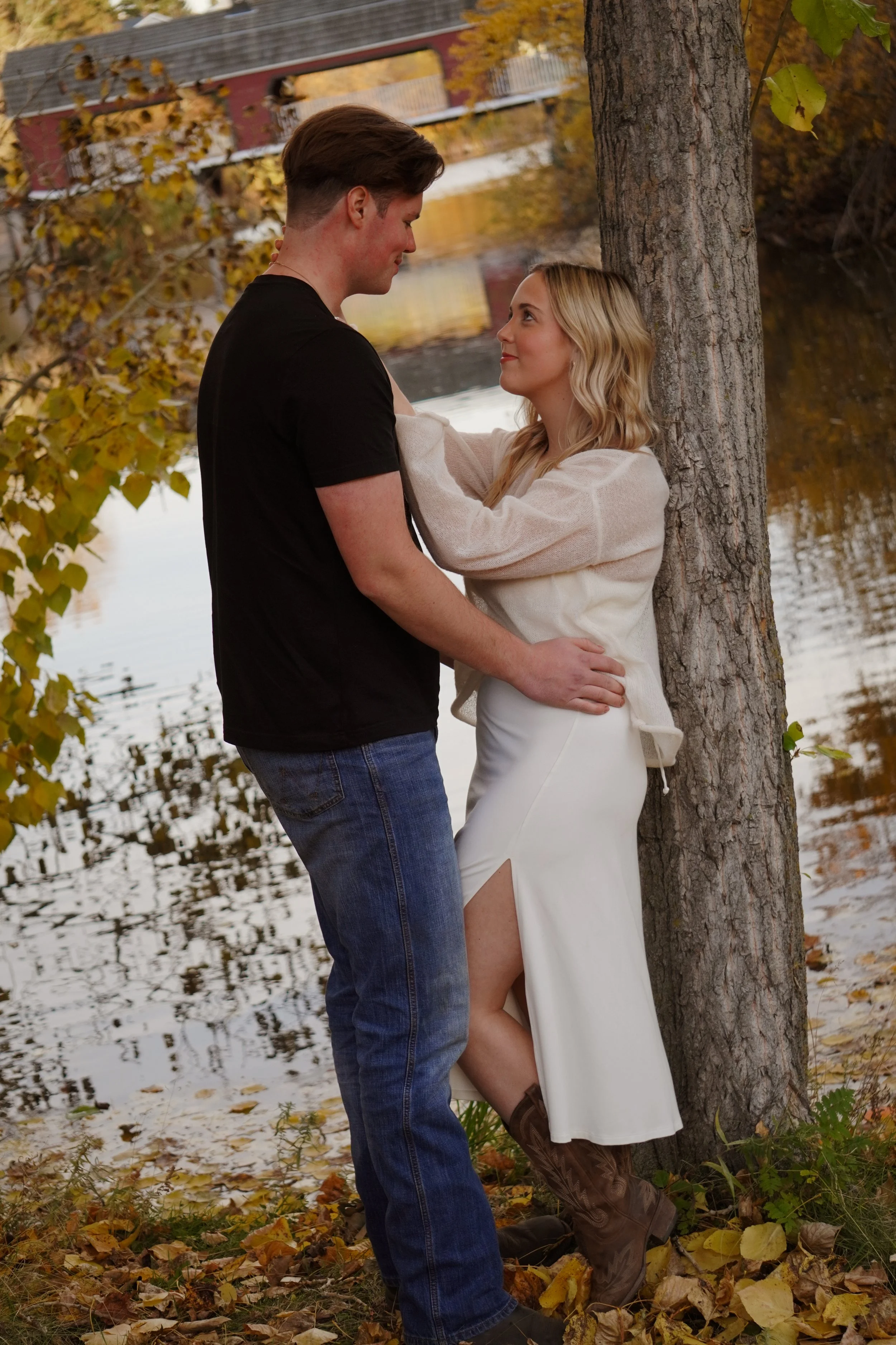 A couple stands close together outdoors near a river, with autumn leaves on the ground and a red bridge in the background. The woman is leaning against a tree, looking up at the man, who is looking down at her. She is wearing a white dress with a slit and brown boots, and he is dressed in a black t-shirt and jeans.