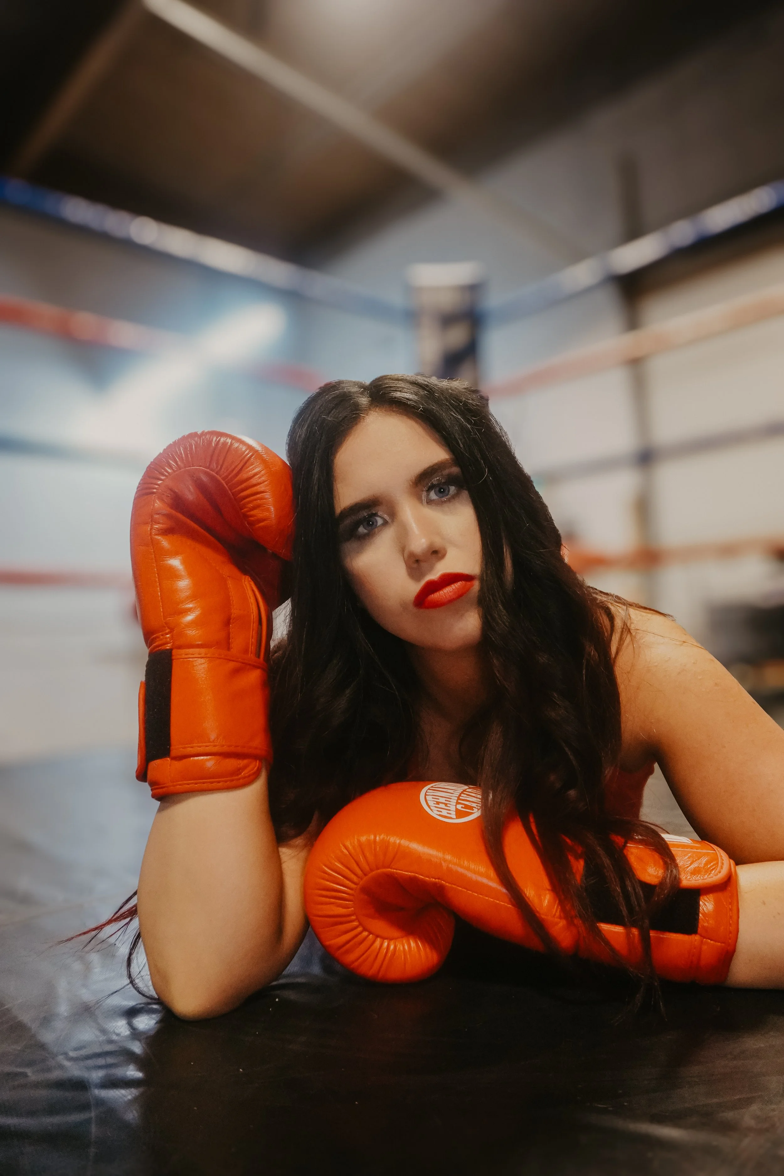 A woman with long dark hair and red lipstick lying on a boxing ring floor, resting her head in her gloved hand and wearing bright orange boxing gloves.