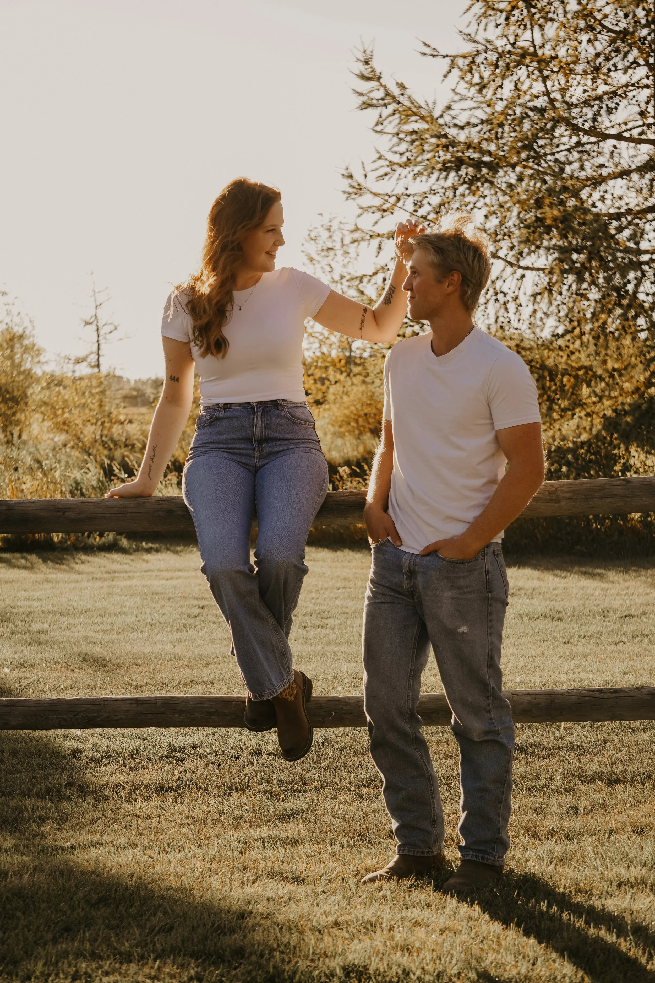 A young woman with long red hair sitting on a wooden fence, casually dressed in a white t-shirt and jeans, smiling and touching the forehead of a young man with blonde hair, wearing a white t-shirt and jeans, standing next to her in an outdoor park during golden hour.