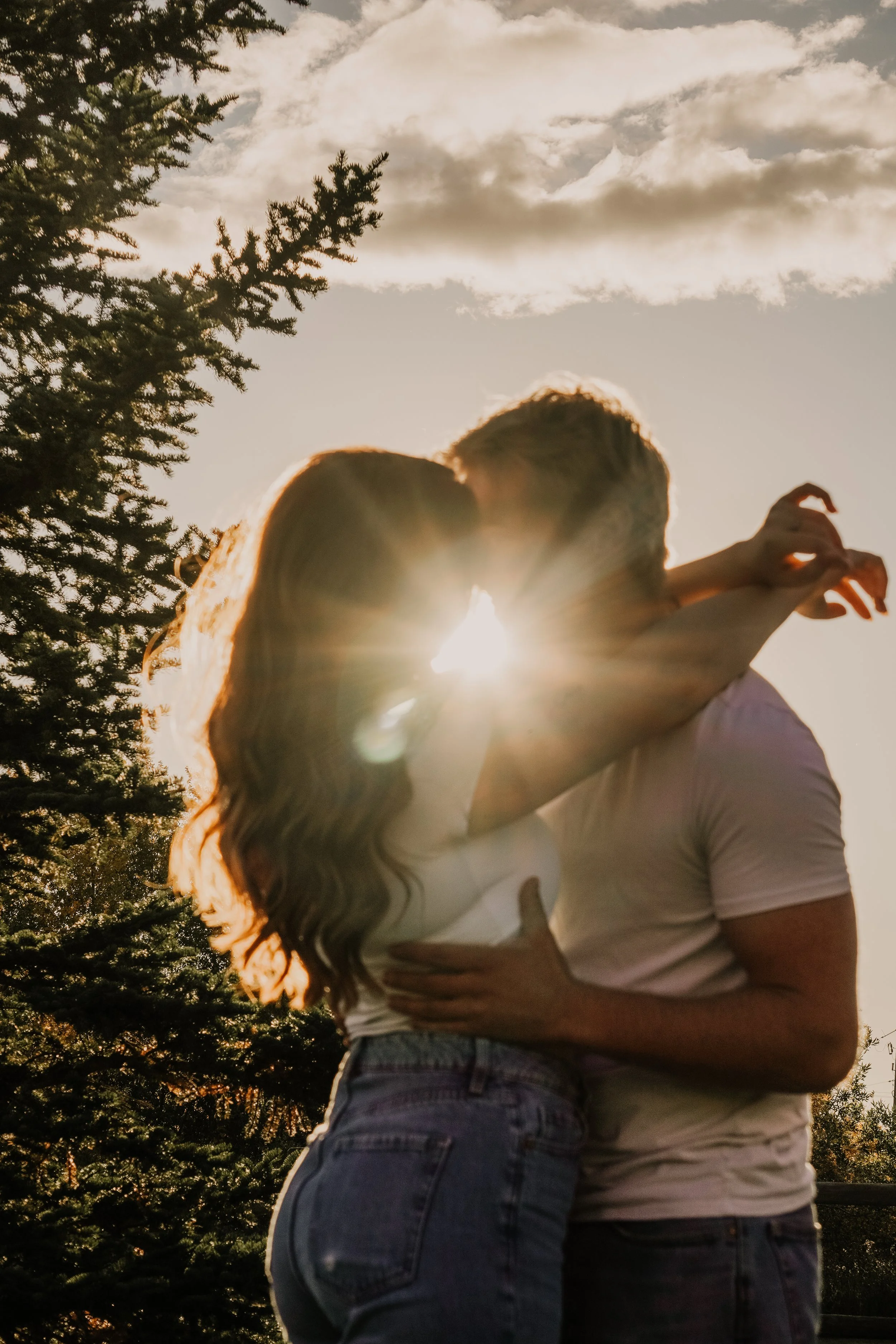 A couple kissing outdoors during sunset with the sun visible between them, trees and cloudy sky in the background.
