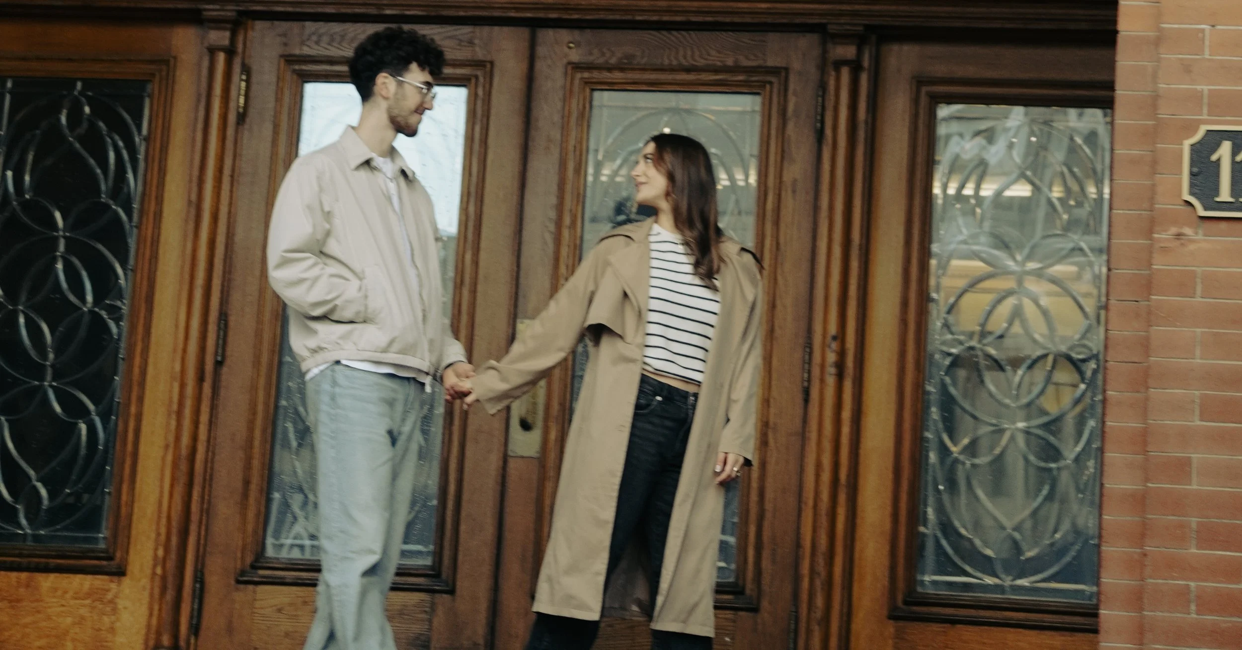 A young man and woman holding hands and talking outside a wooden front door of a house. The man has curly dark hair, glasses, and is wearing a beige jacket and light-colored pants. The woman has long brown hair, is wearing a beige trench coat, a striped shirt, and black pants.